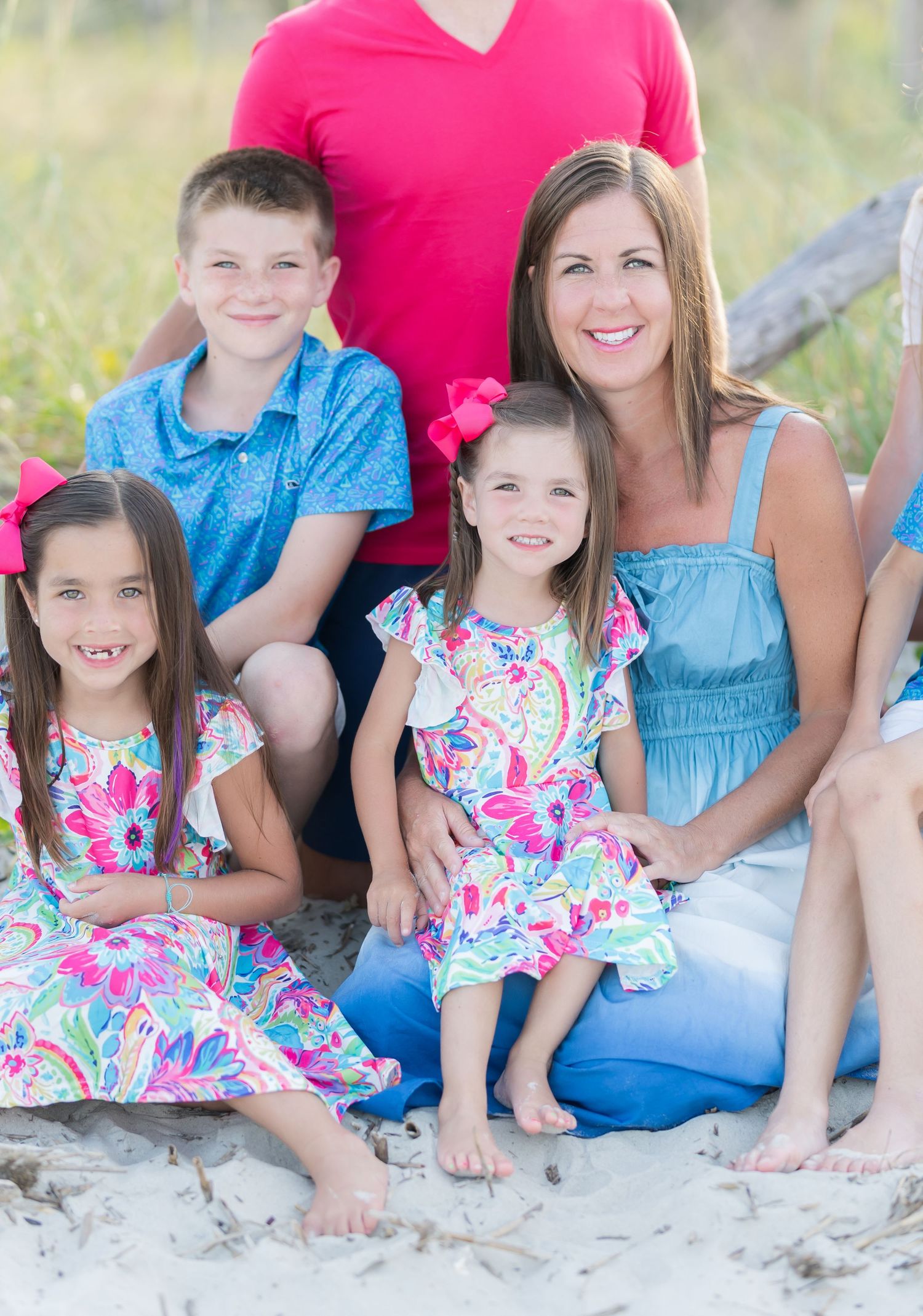 Family in coordinated outfits sitting together on sandy beach with pink floral dresses and blue clothing.