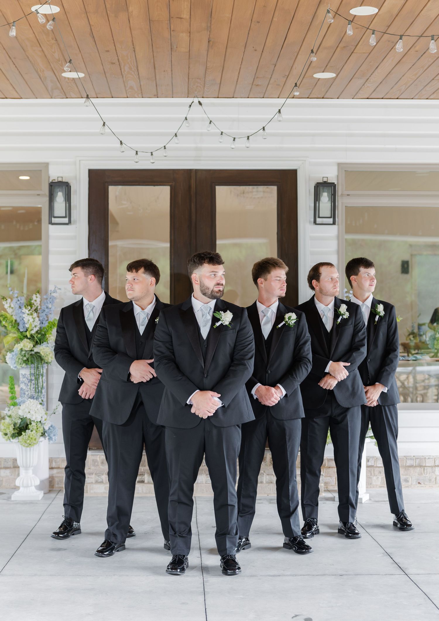 Groom in black suit stands with groomsmen in front of a white wedding venue entrance.