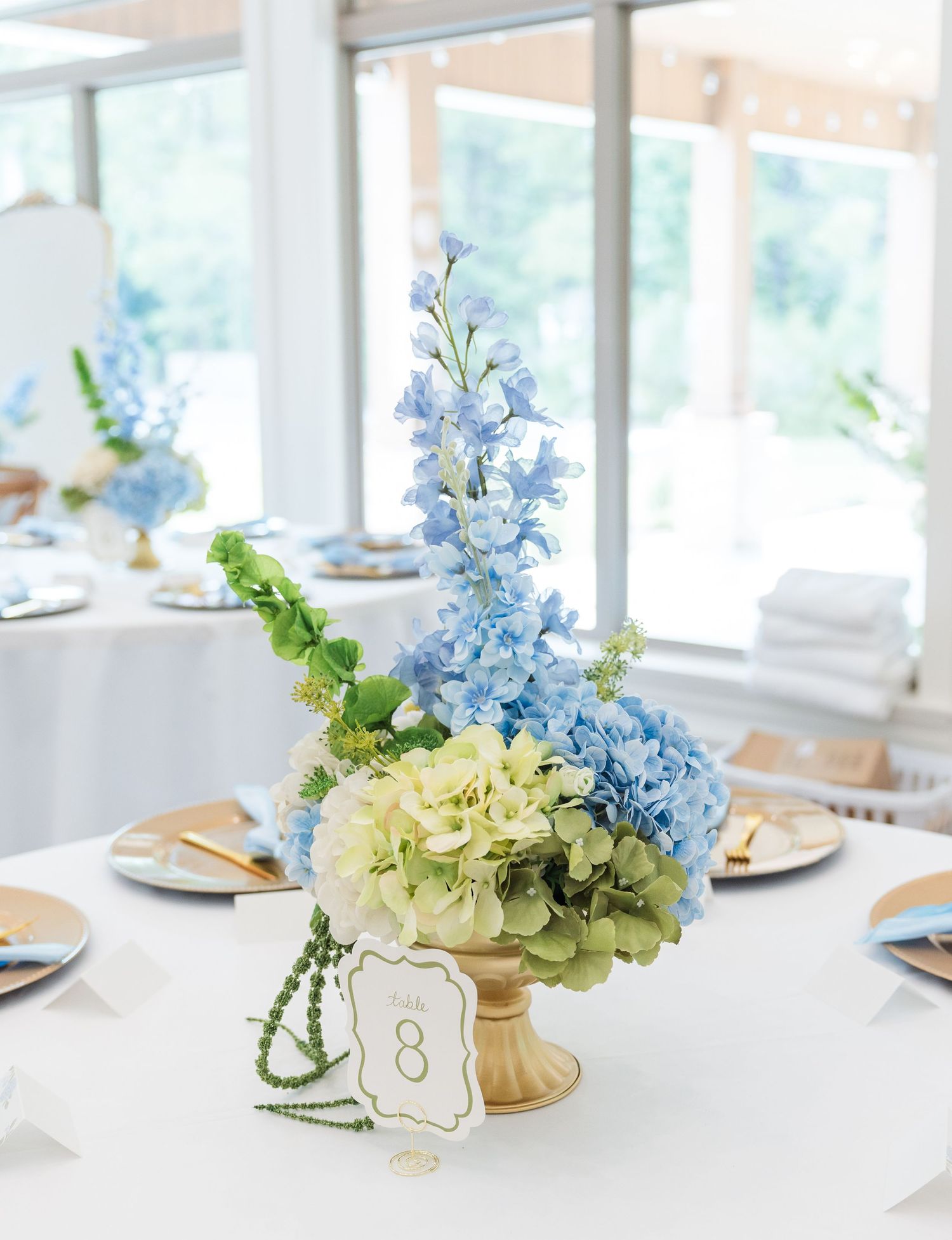 Blue delphinium and green hydrangea wedding centerpiece in a gold pedestal vase with table number 8.