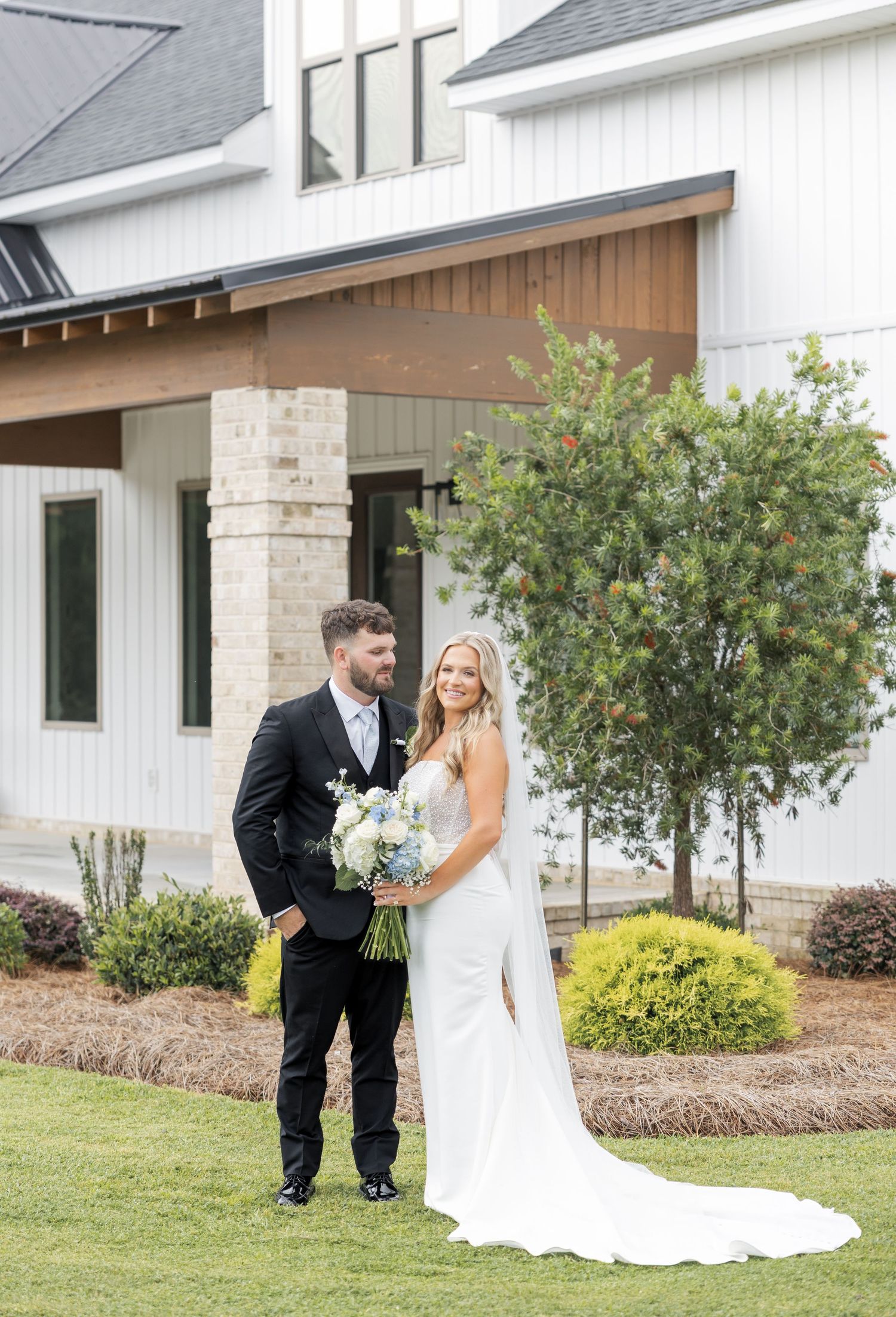 Bride and groom posing outside modern white farmhouse wedding venue with blue hydrangea bouquet.
