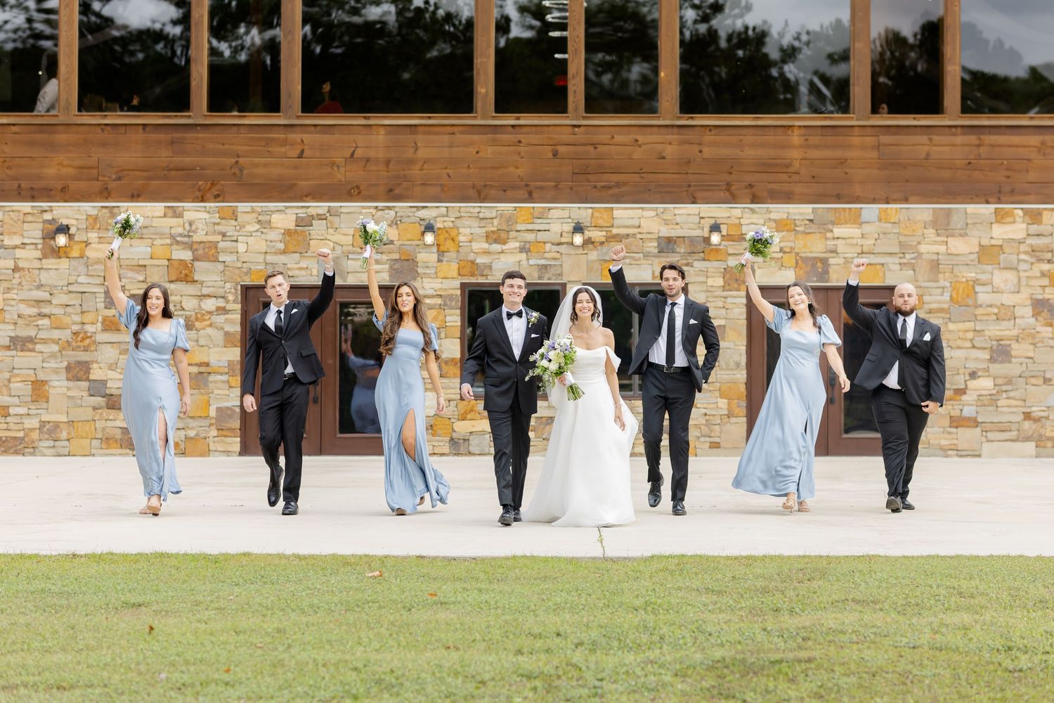 Wedding party members in formal attire pose in a line against a brick wall and grassy lawn.