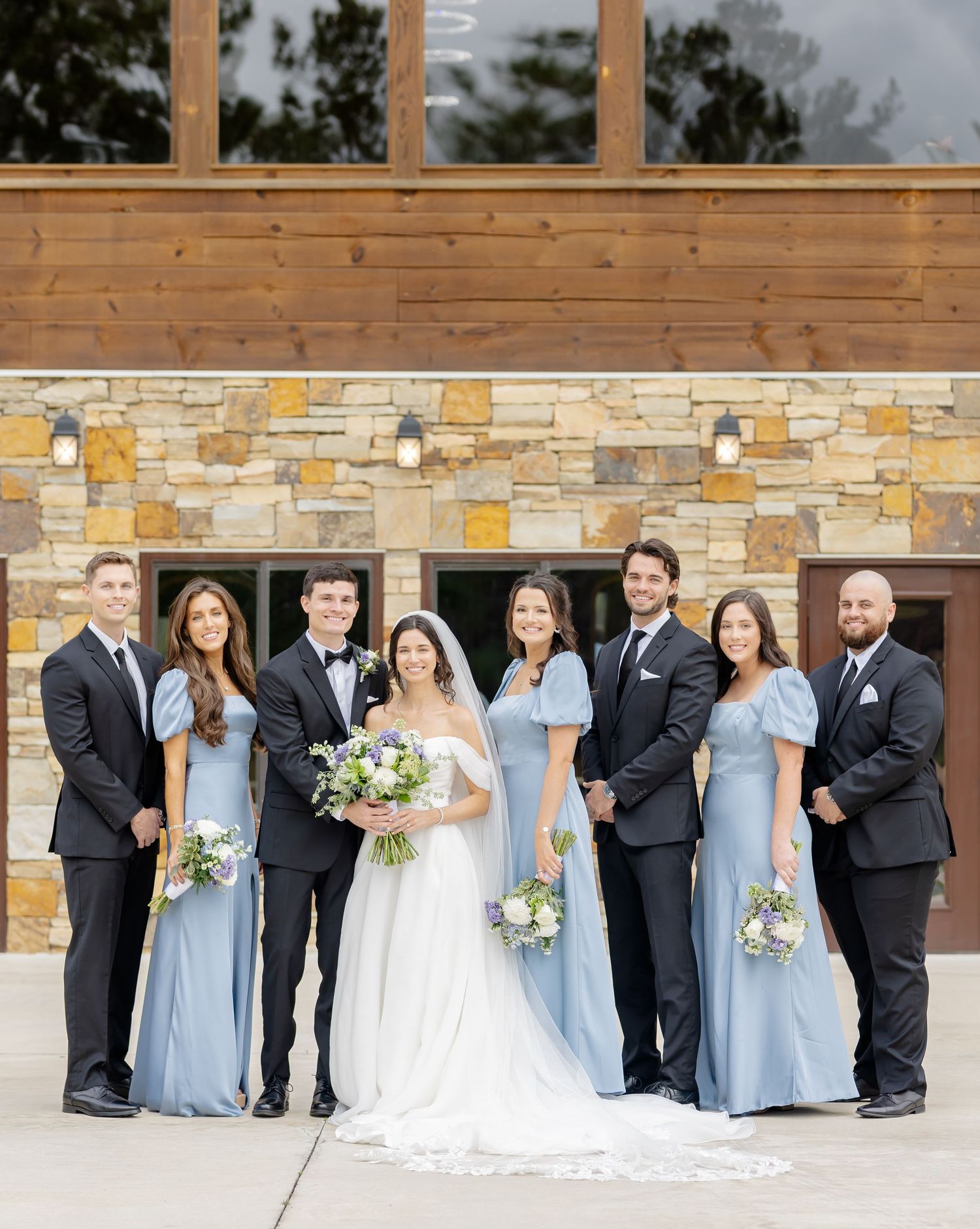 Wedding party poses in formal attire against modern stone and wood building exterior.