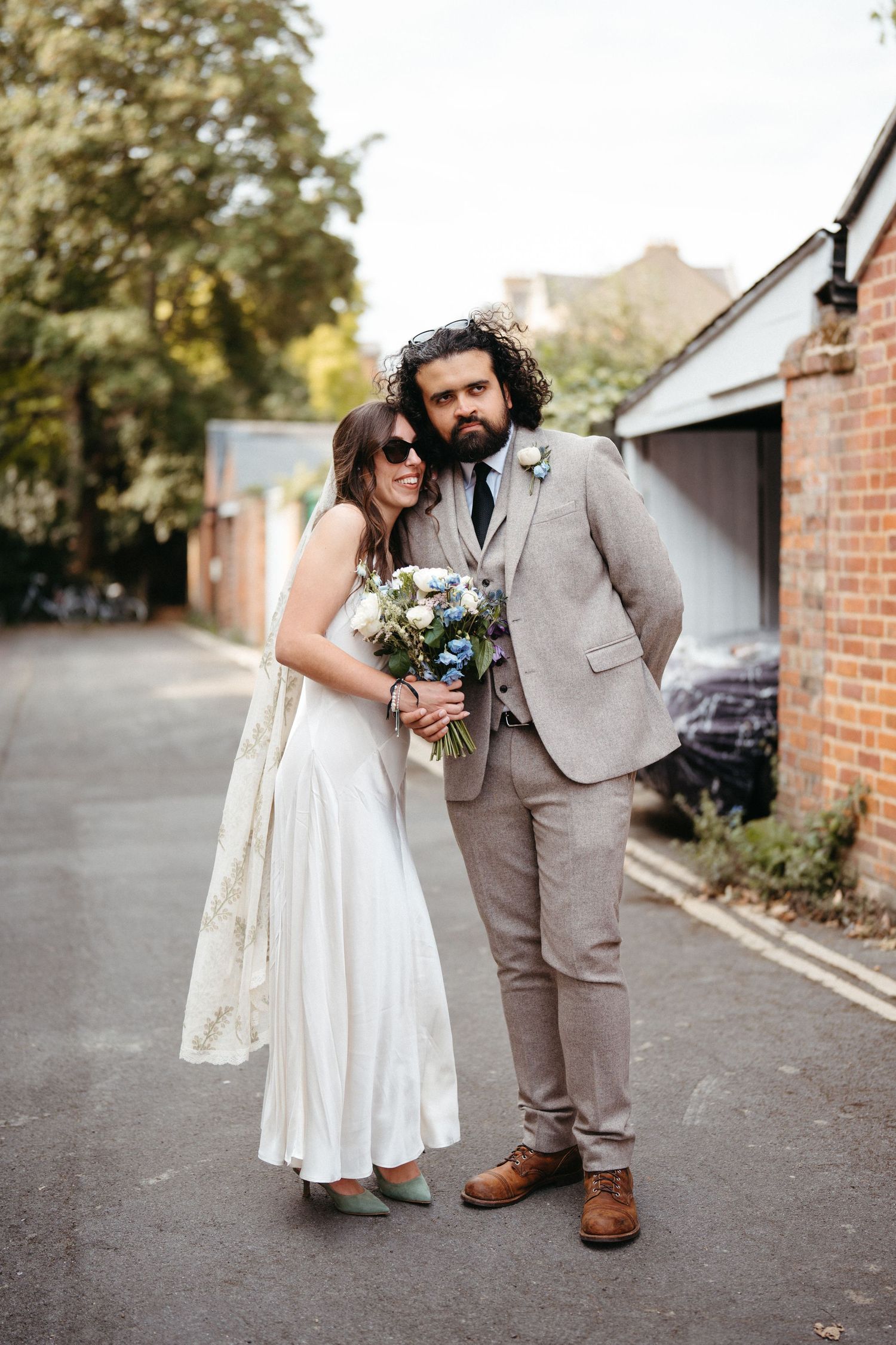 Wedding couple poses together outside brick building, bride in white dress holding bouquet and groom in tan suit.