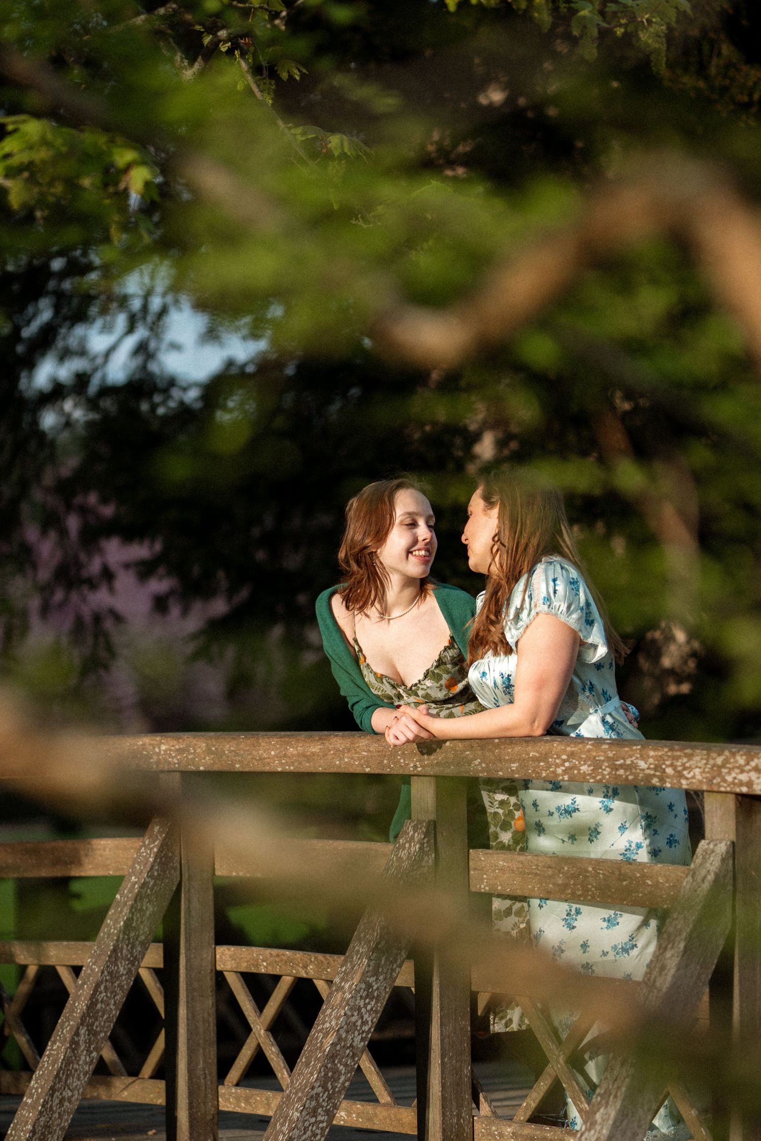 Two people share a close conversation while leaning on a wooden bridge railing on a sunny afternoon. Oxford wedding photographer