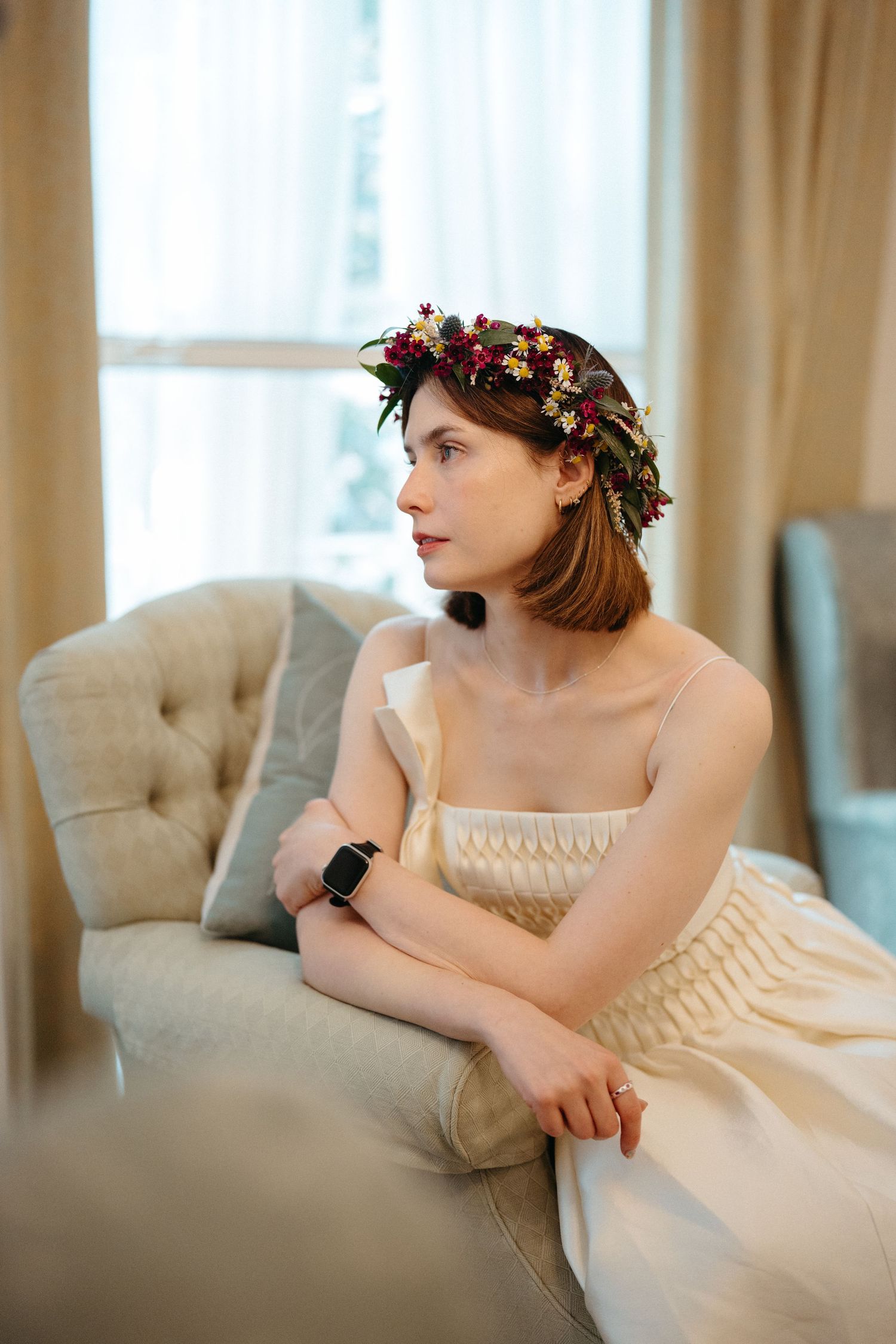 Beautiful floral crown adorns vintage-inspired cream dress as figure reclines on elegant tufted gray chair near window.