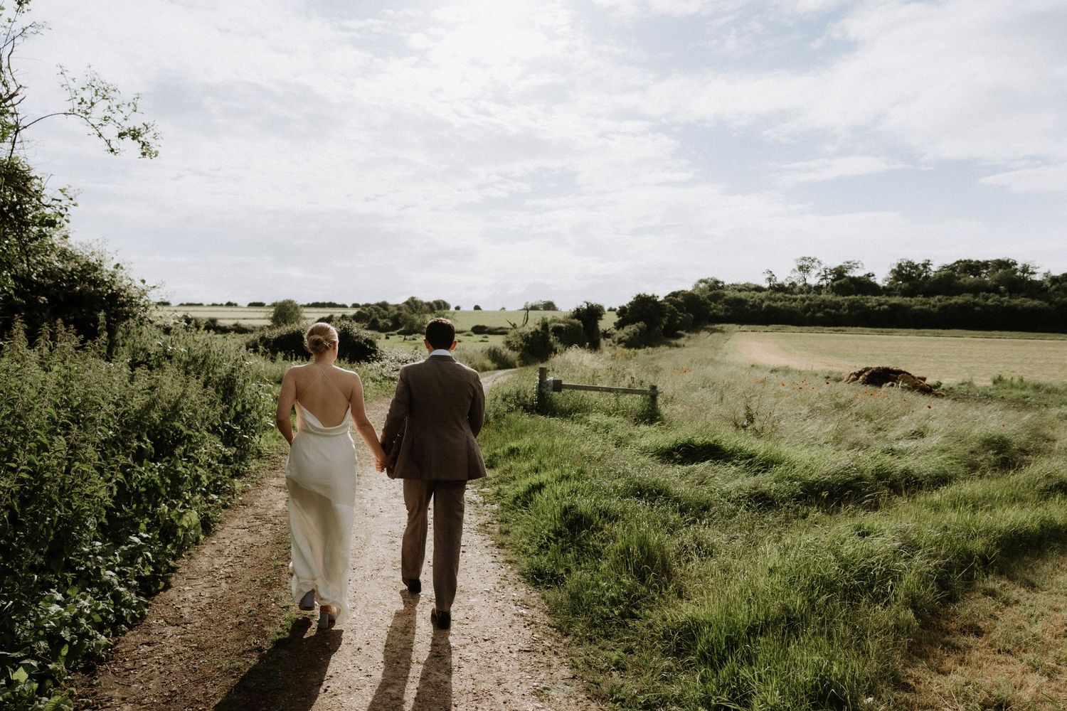 A couple walks hand in hand down a scenic countryside path surrounded by green fields and rolling hills.