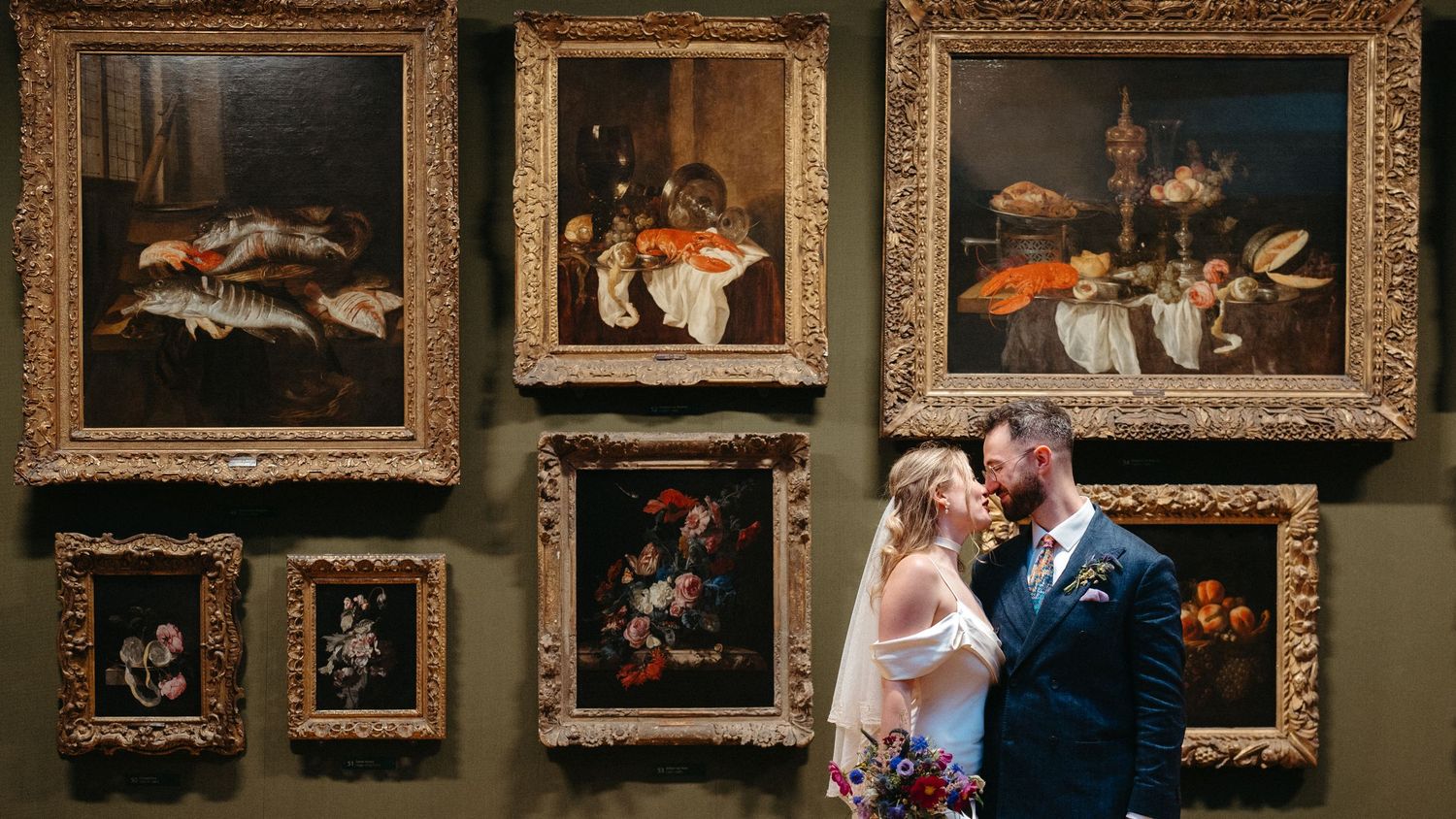 A couple embraces in front of a gallery wall displaying ornate framed still life paintings of food and table settings.