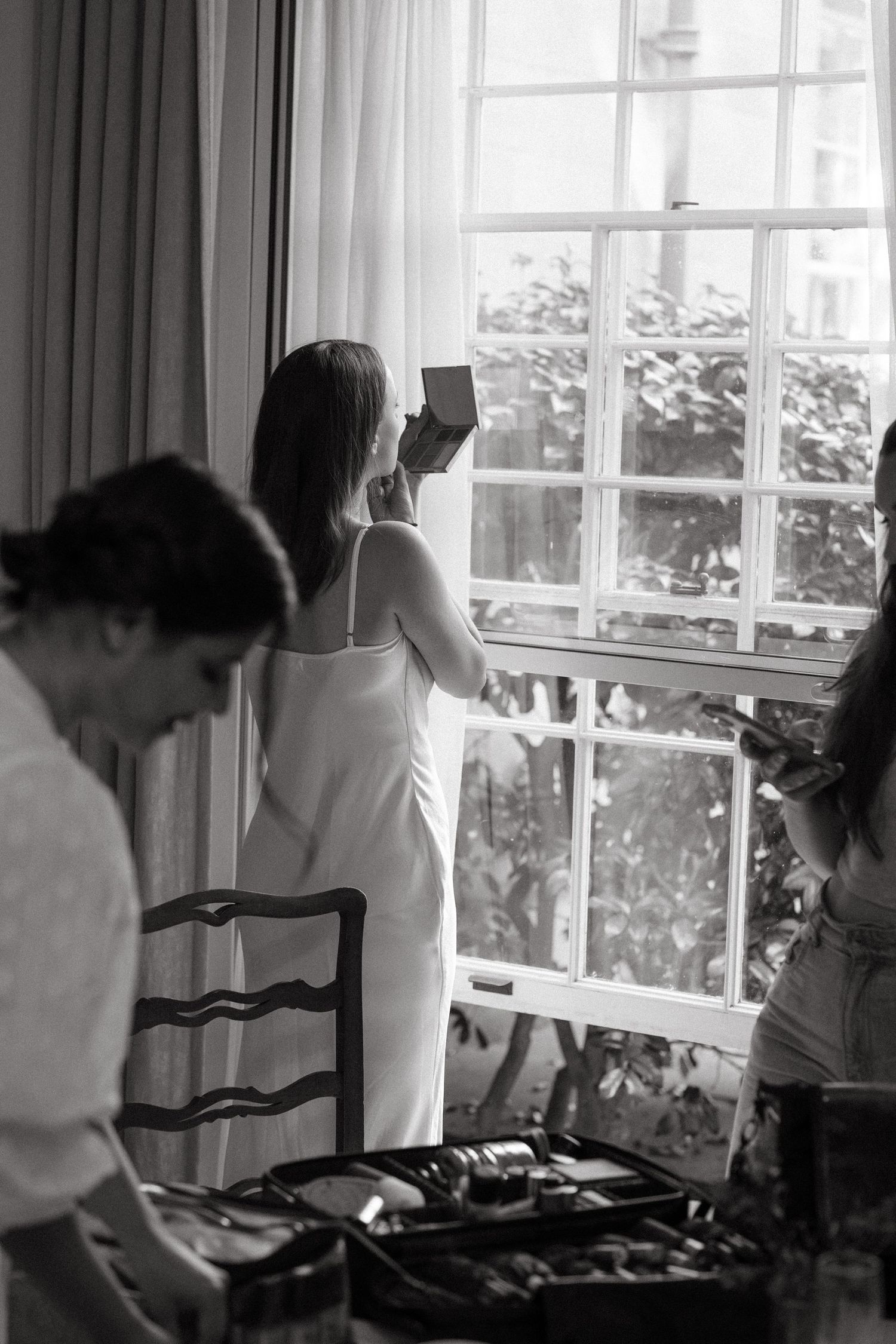 Black and white photo showing people in a bright room with large windows, someone in white dress looking outside.
