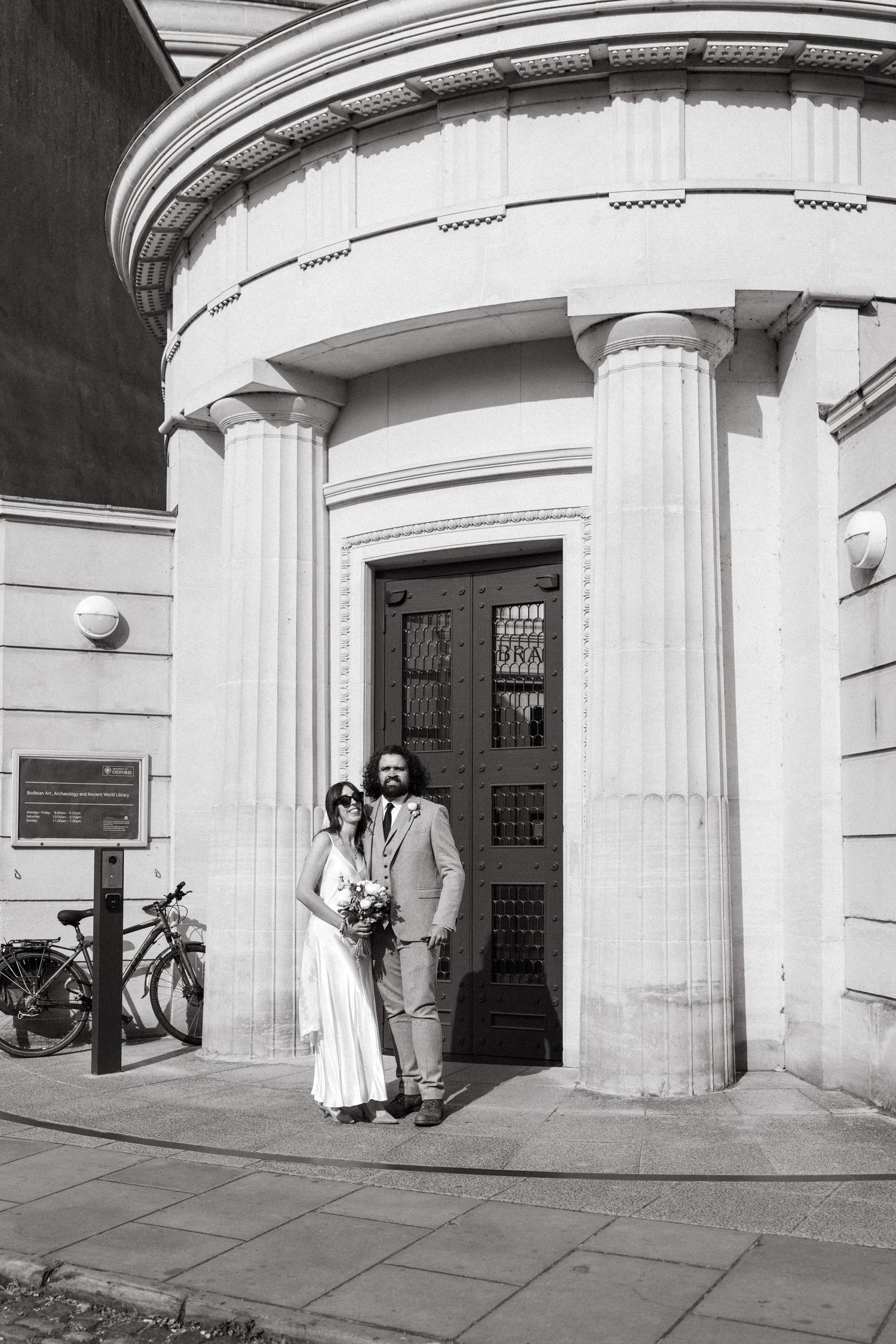A couple poses by a neoclassical building entrance with white columns and a curved facade in black and white photography.
