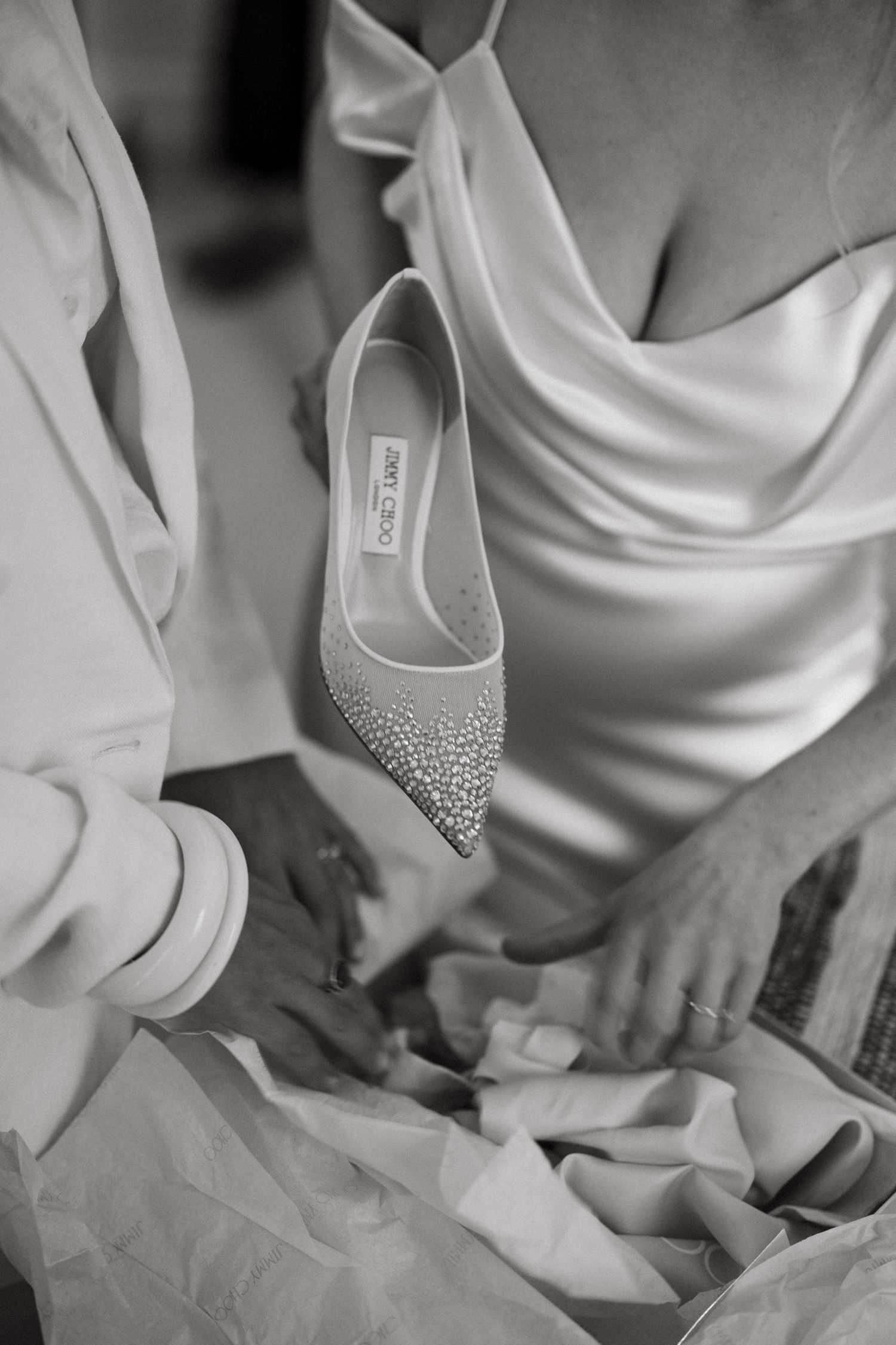 A sparkly rhinestone-encrusted wedding heel displayed in black and white against a silky dress background.