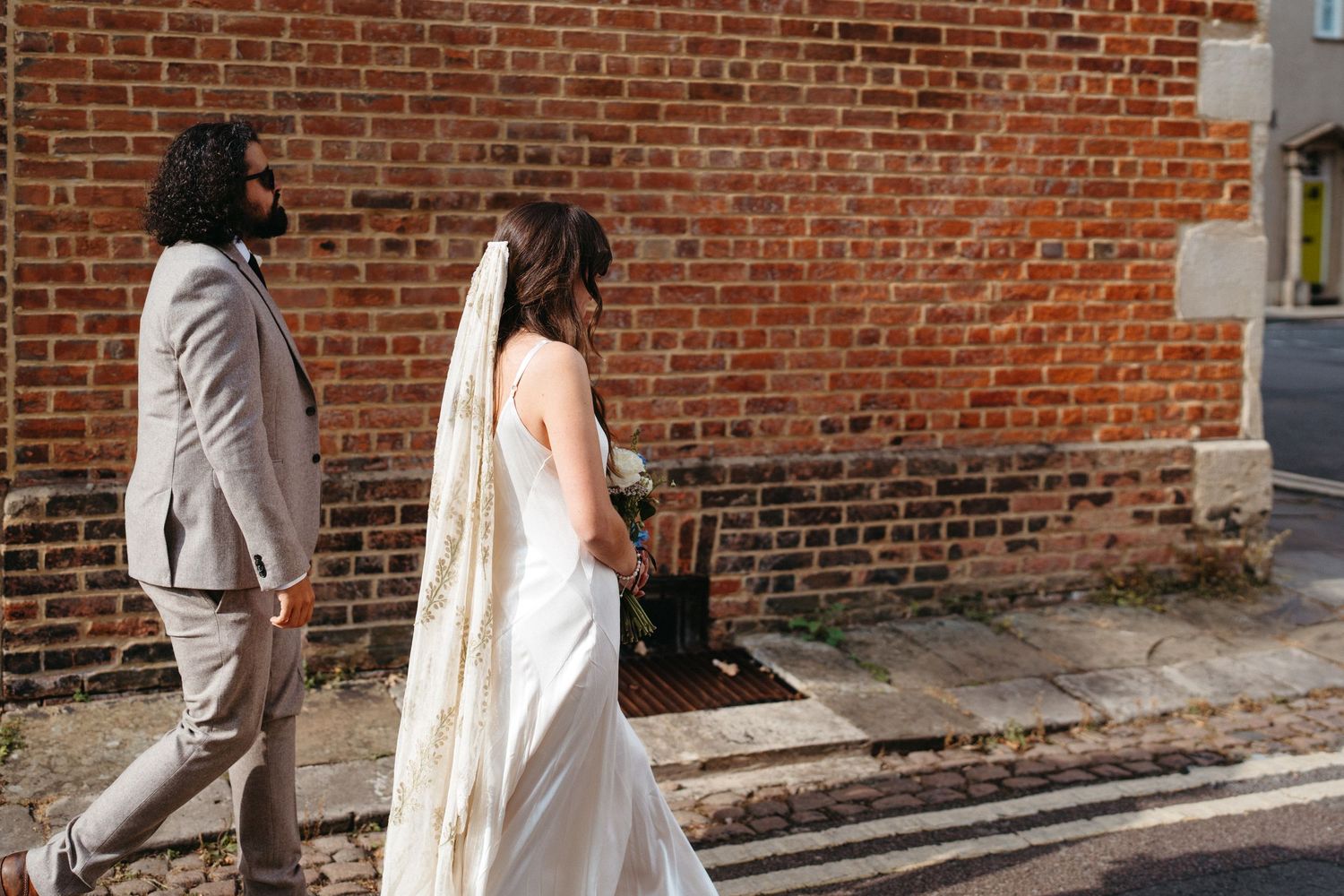 Two people walking along a brick wall wearing neutral colored outfits in an urban street setting.
