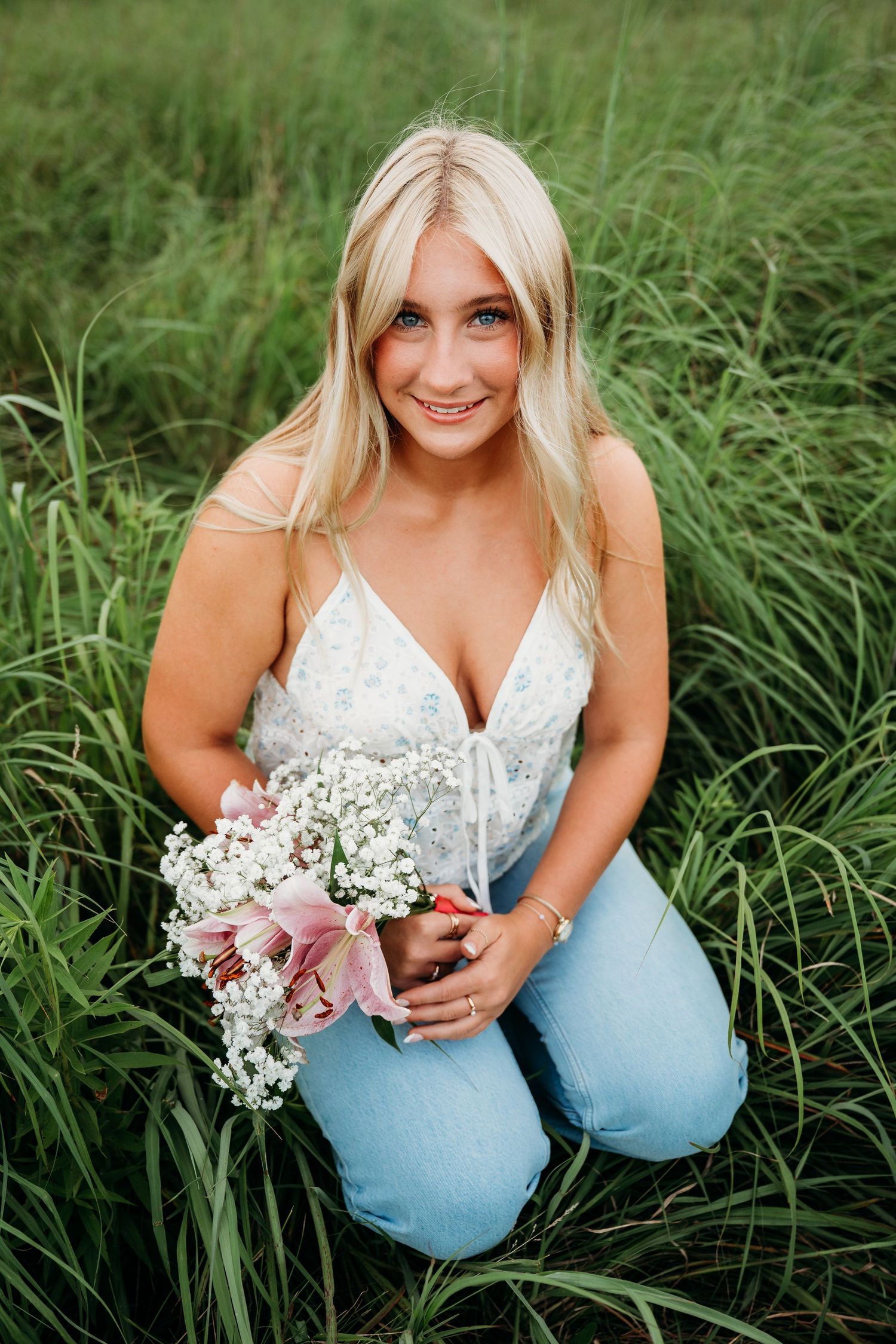 Young woman in white dress crouches in tall grass holding delicate wildflowers in a summer photo series.