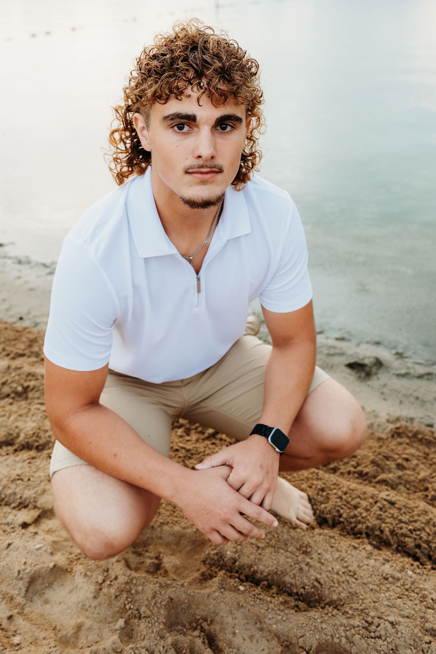 A person in a white polo shirt and khaki shorts kneels on sandy terrain against a misty natural backdrop.
