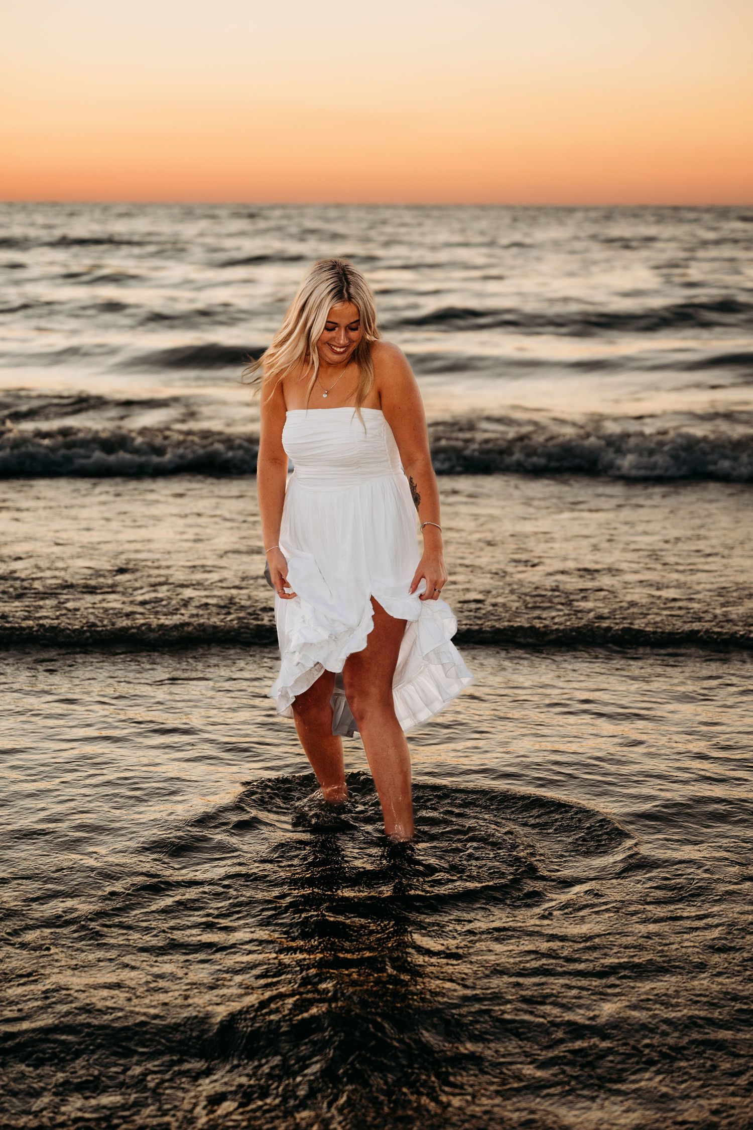 A person in a flowing white sundress stands in shallow ocean waters at sunset, creating a serene coastal scene.