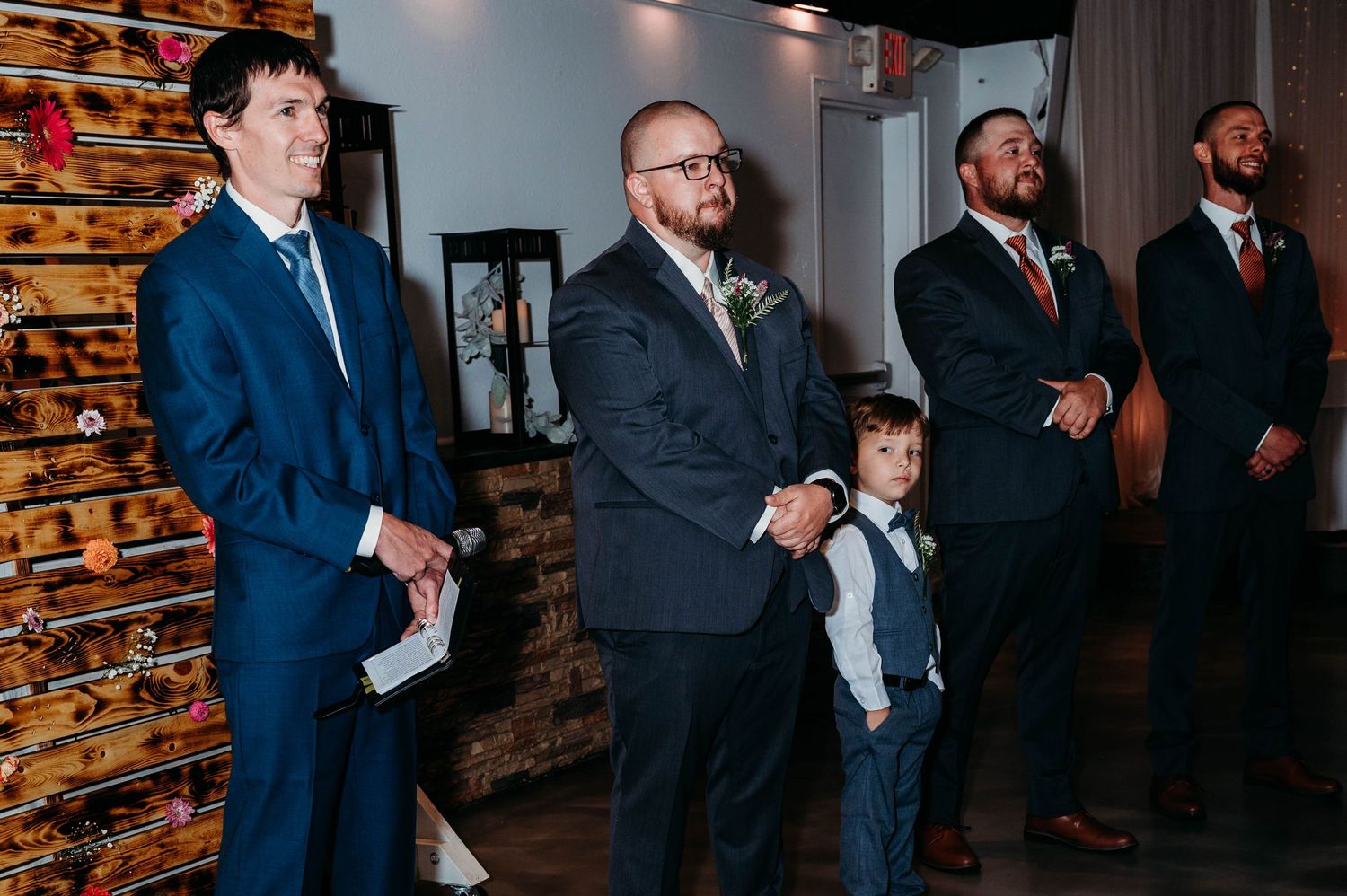Wedding party members in formal attire gather in an indoor venue with warm lighting and decorative elements.