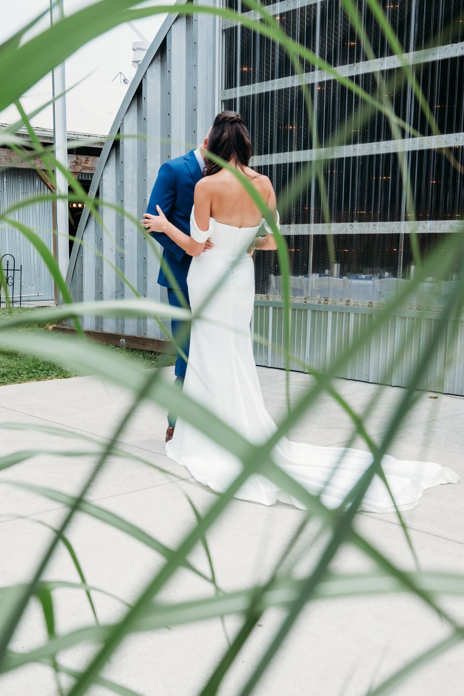 Through green leaves, a couple in wedding attire embraces outside a modern building with glass panels.