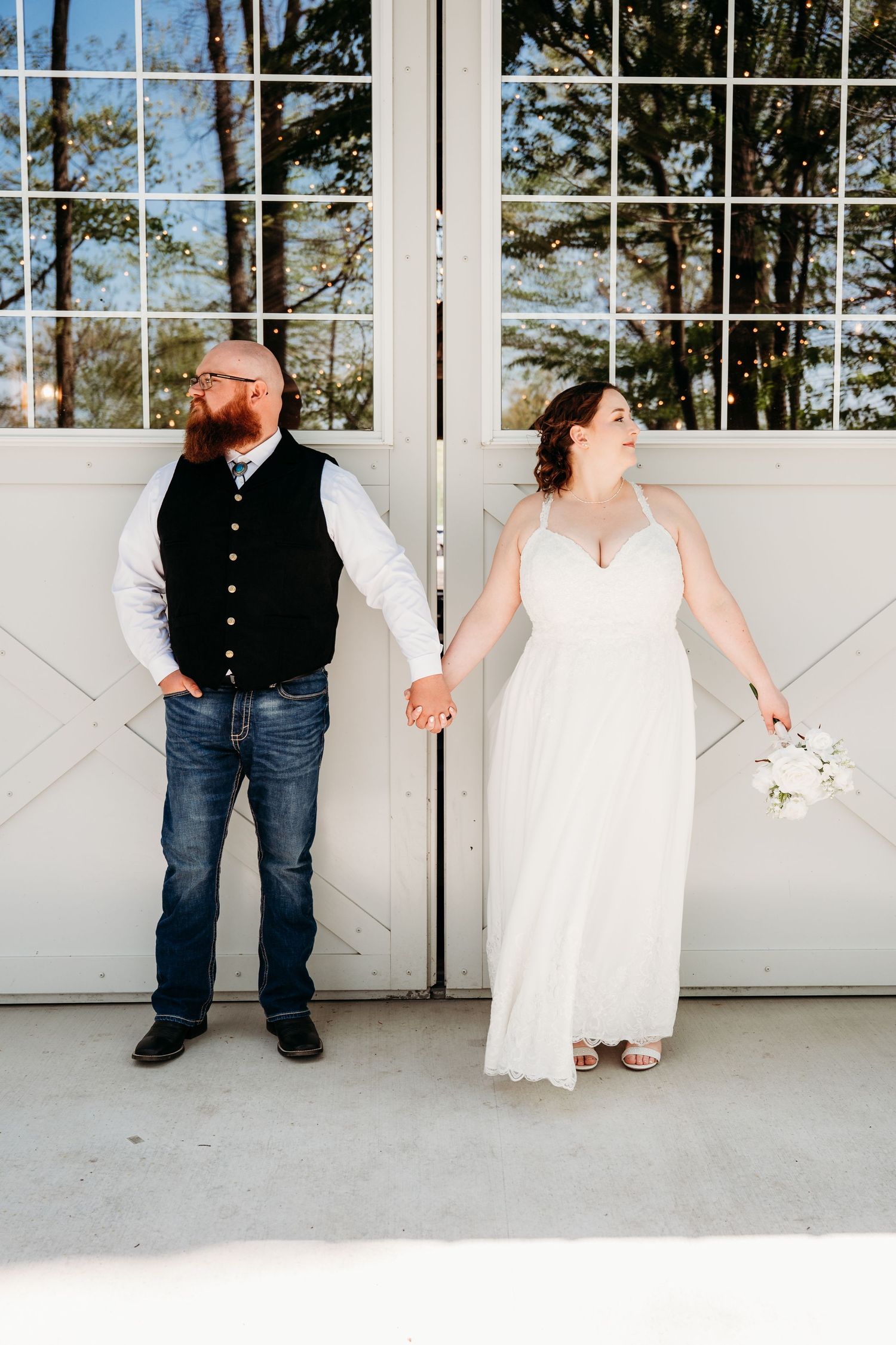 A couple poses in front of white barn doors, with the bride in a flowing white dress and groom in a vest and jeans.