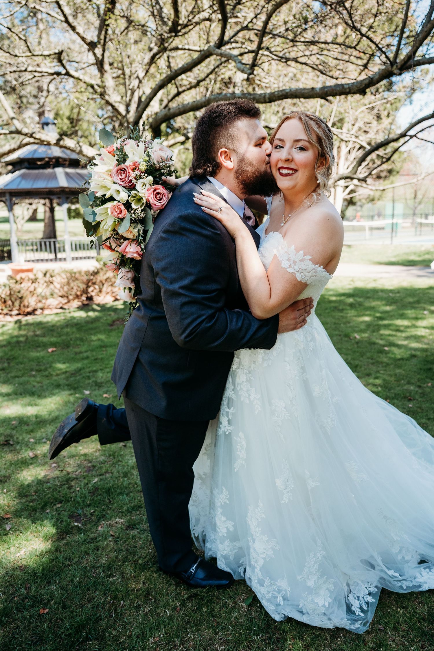 A joyful moment captured outdoors with a couple in wedding attire embracing under a blooming tree on a sunny day.