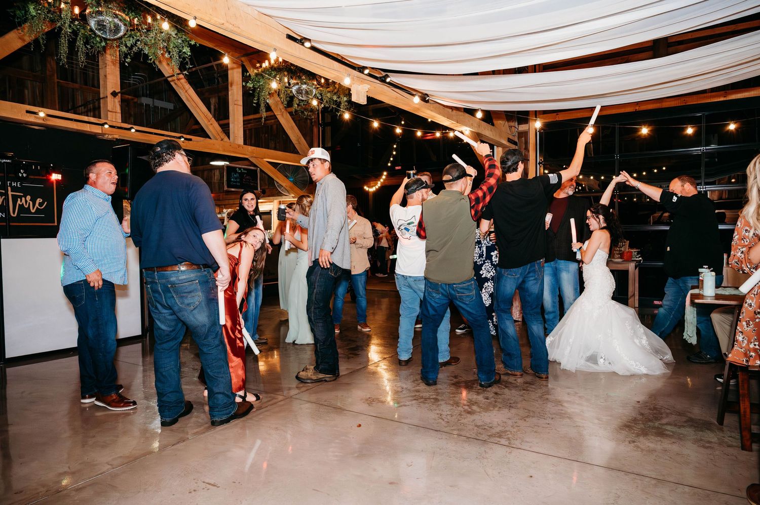 Wedding reception party with guests dancing under string lights in a rustic barn venue.