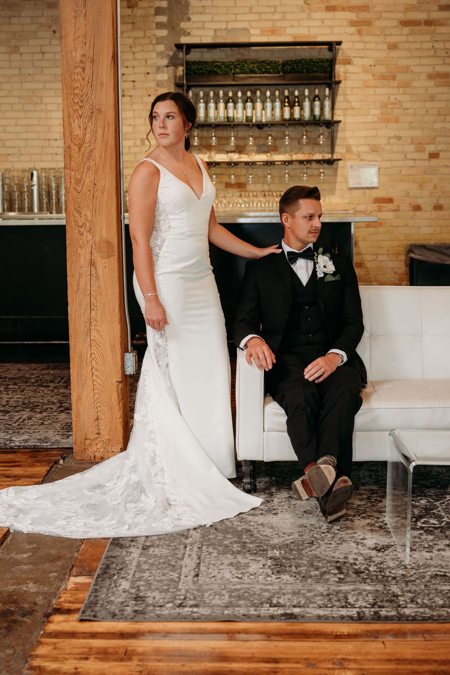 A bride in a white lace gown and groom in a black suit sit together on a modern couch in a rustic brick venue.