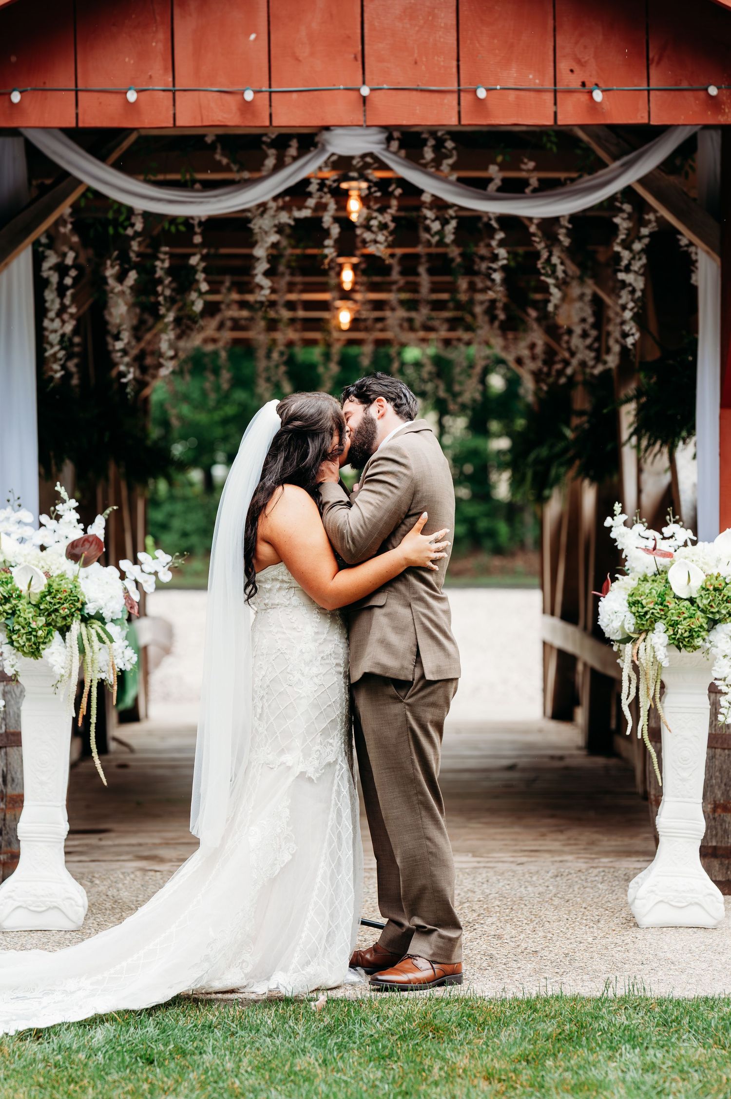 A bride and groom share their first kiss at a rustic barn wedding ceremony, surrounded by white floral arrangements.