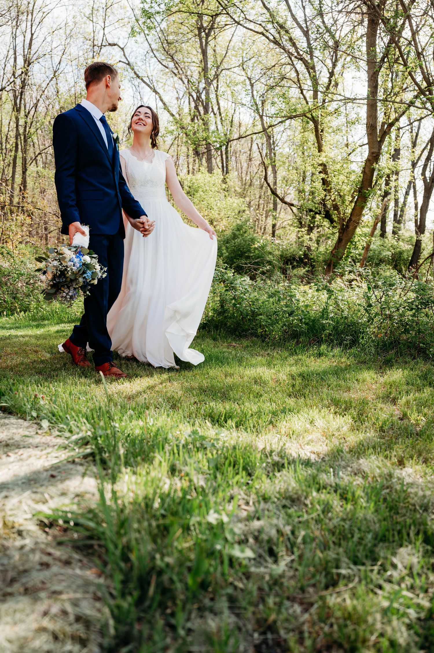 A bride and groom walk hand-in-hand through a lush green forest, she lifts her white gown while he holds a bouquet.