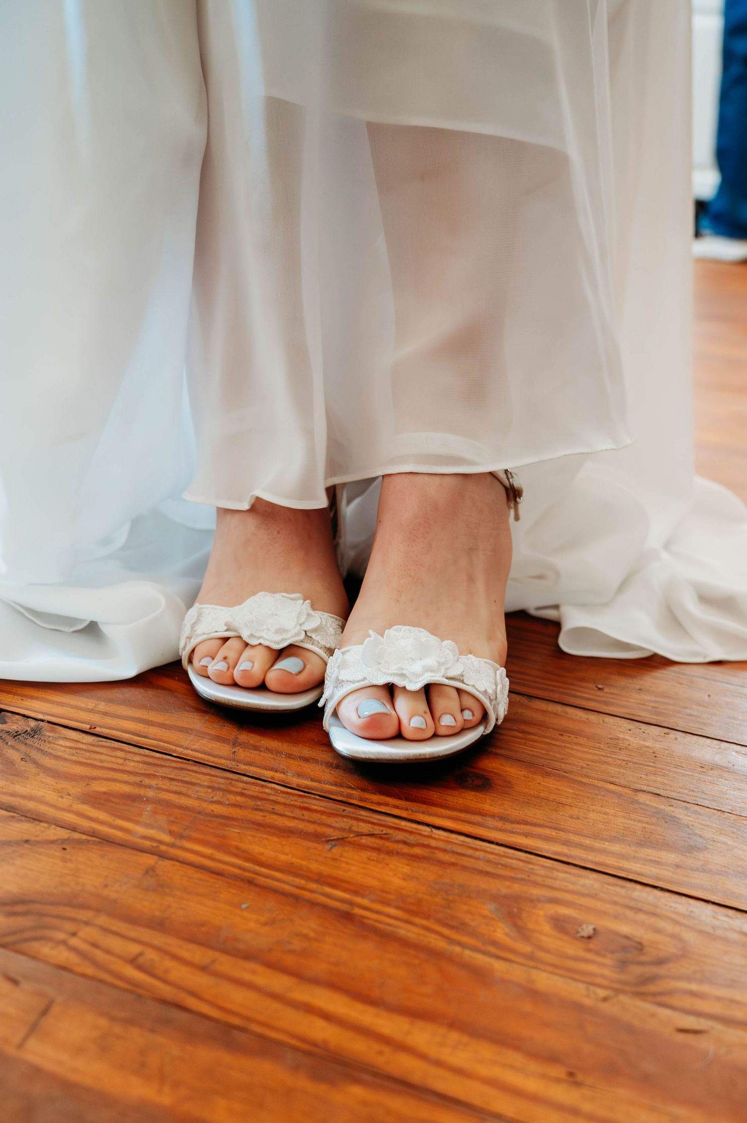 Bride wearing white floral sandals with blue toenails, standing on rustic hardwood floor in a flowing white wedding gown.