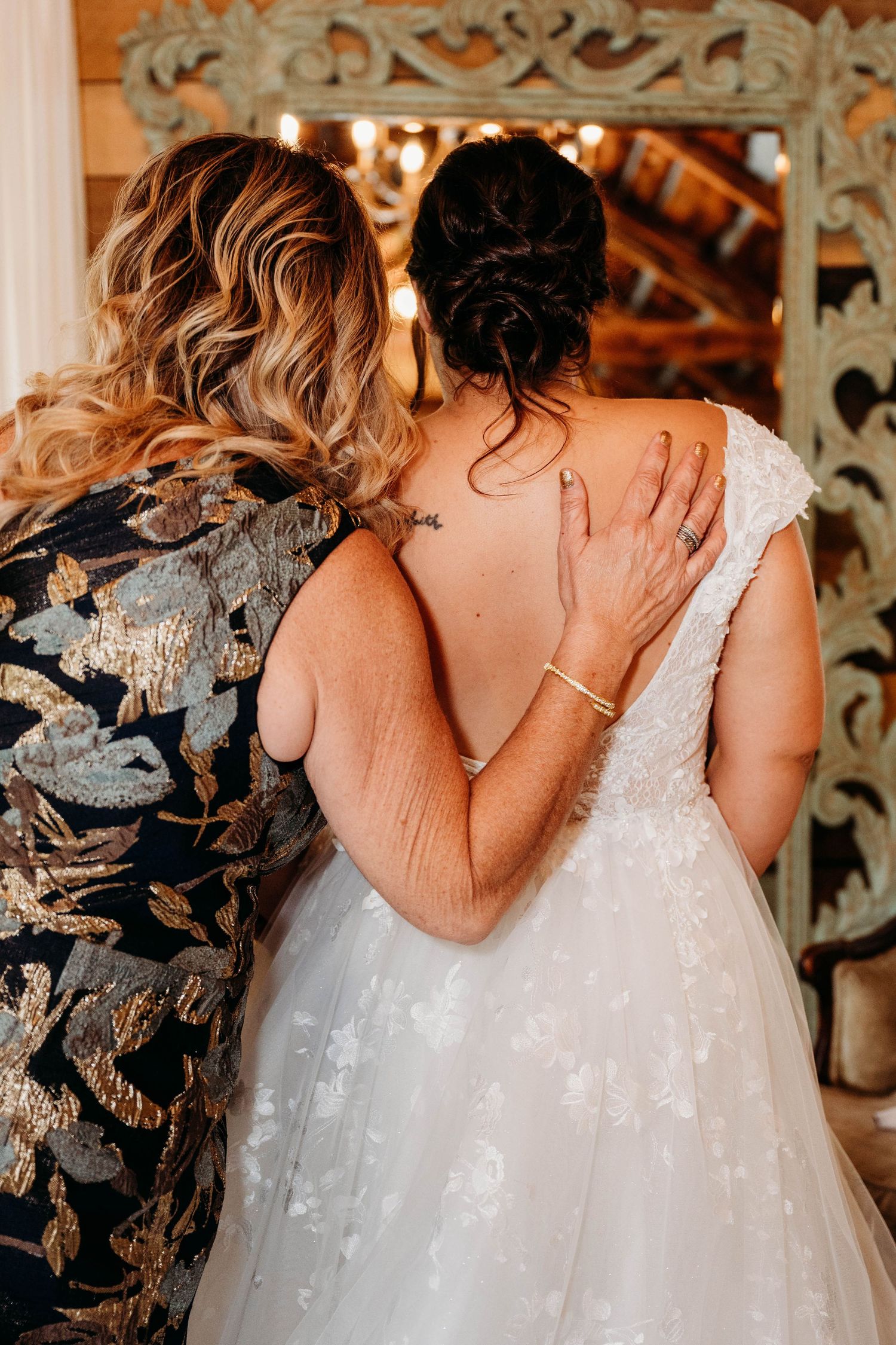 Two people embrace in formal attire, one wearing a floral brocade dress and one in a white lace wedding gown.