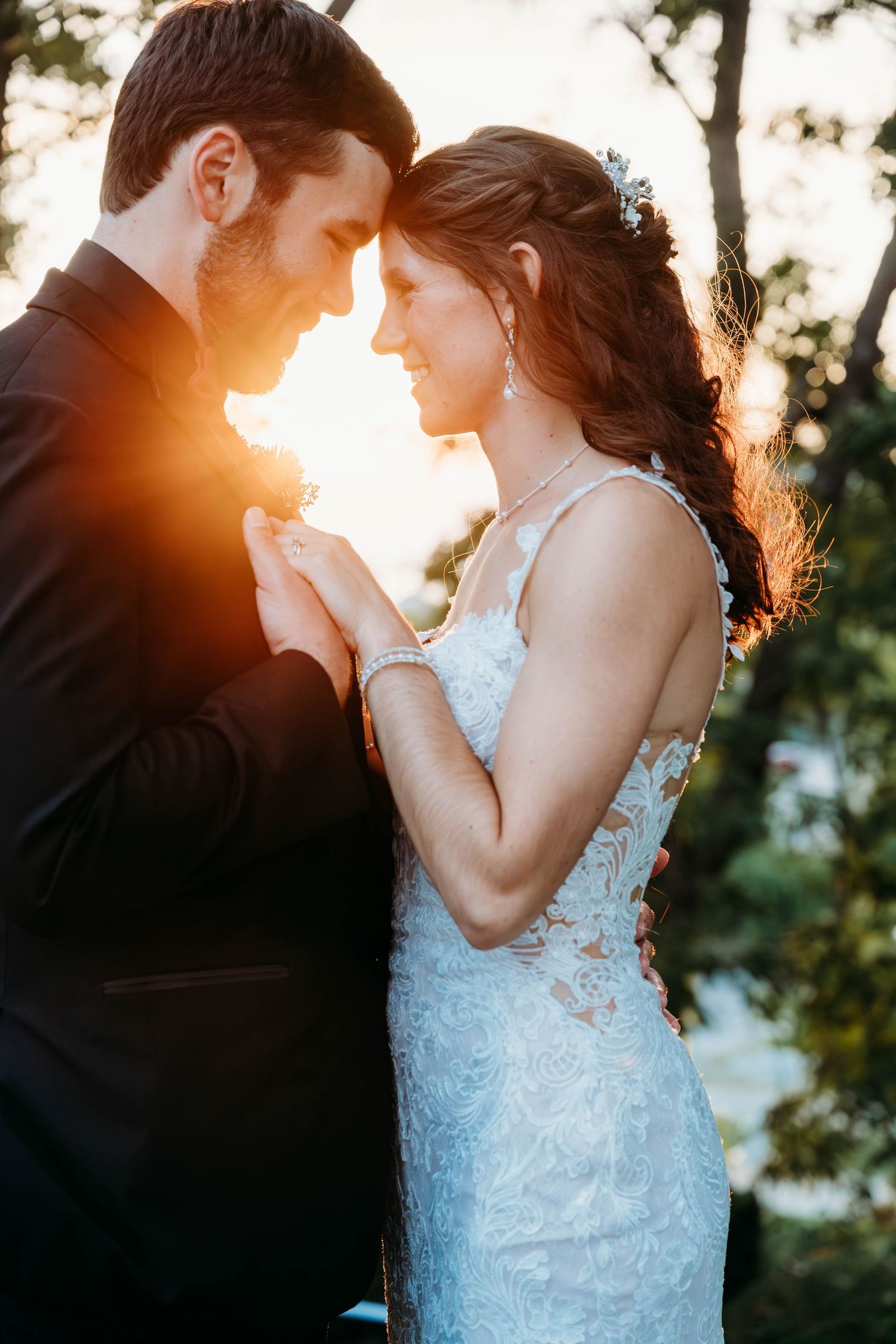 A couple in wedding attire embraces during golden hour sunset, with sunlight creating a warm backlit glow through trees.
