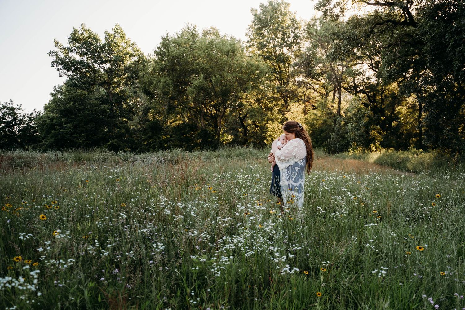 Two people embrace in a sunlit wildflower meadow at golden hour with trees in the background.