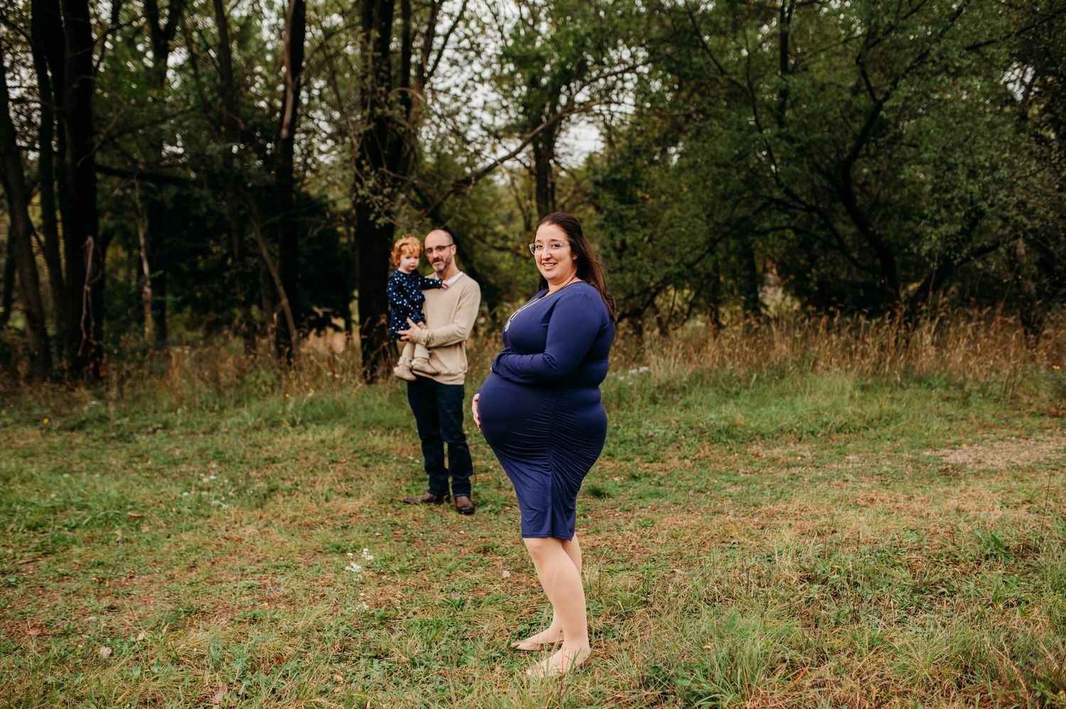Family outdoor photo session in a natural wooded setting with tall trees and autumn grass in the background.