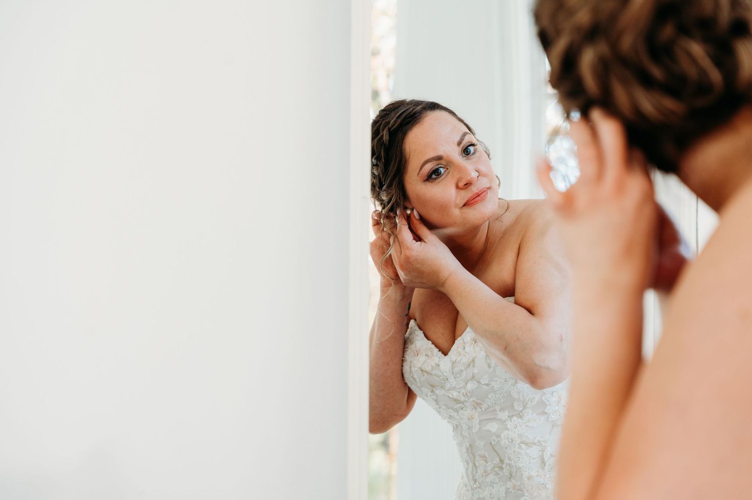 A bride in a white lace dress adjusts her hair while looking in a mirror during wedding preparations.