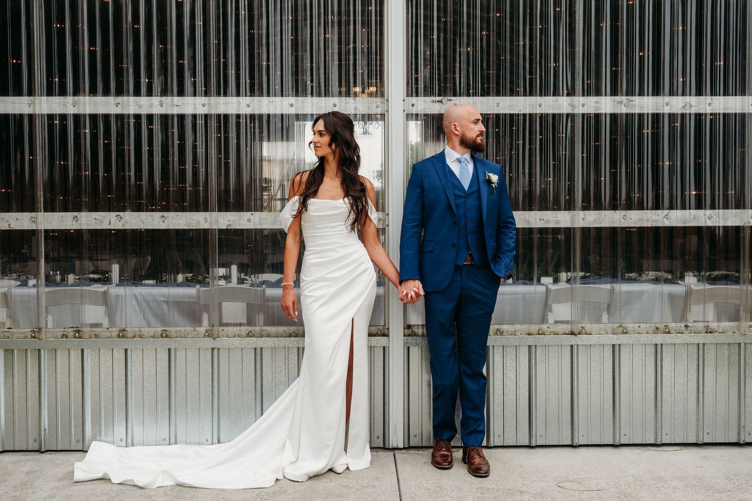 Bride in white gown and groom in navy suit hold hands outdoors against a modern glass building backdrop.