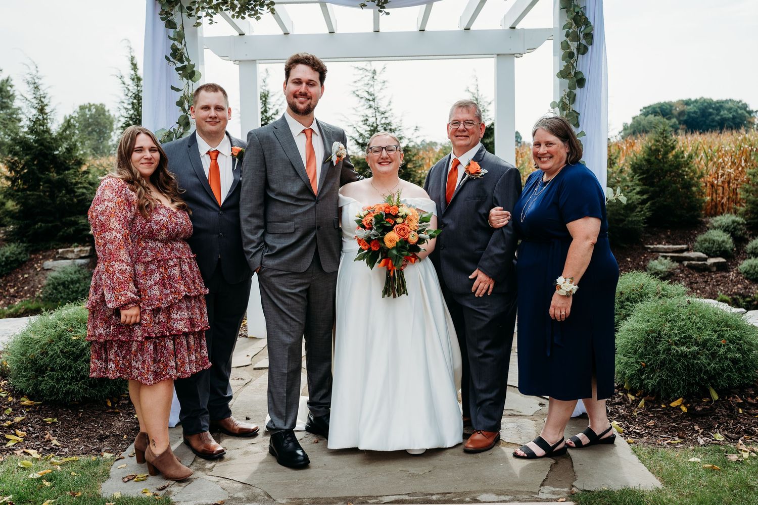 Wedding party of six smiling outdoors in a garden, bride in white gown holding colorful bouquet, guests in formal attire.