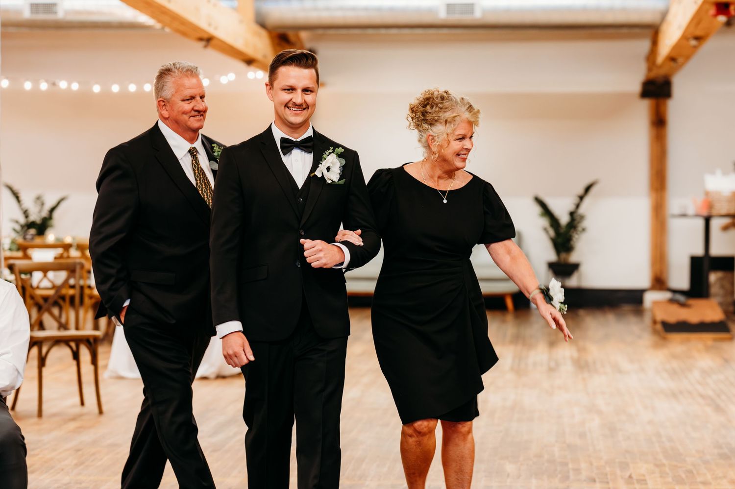 Groom in black tuxedo escorted by parents during wedding reception entrance.