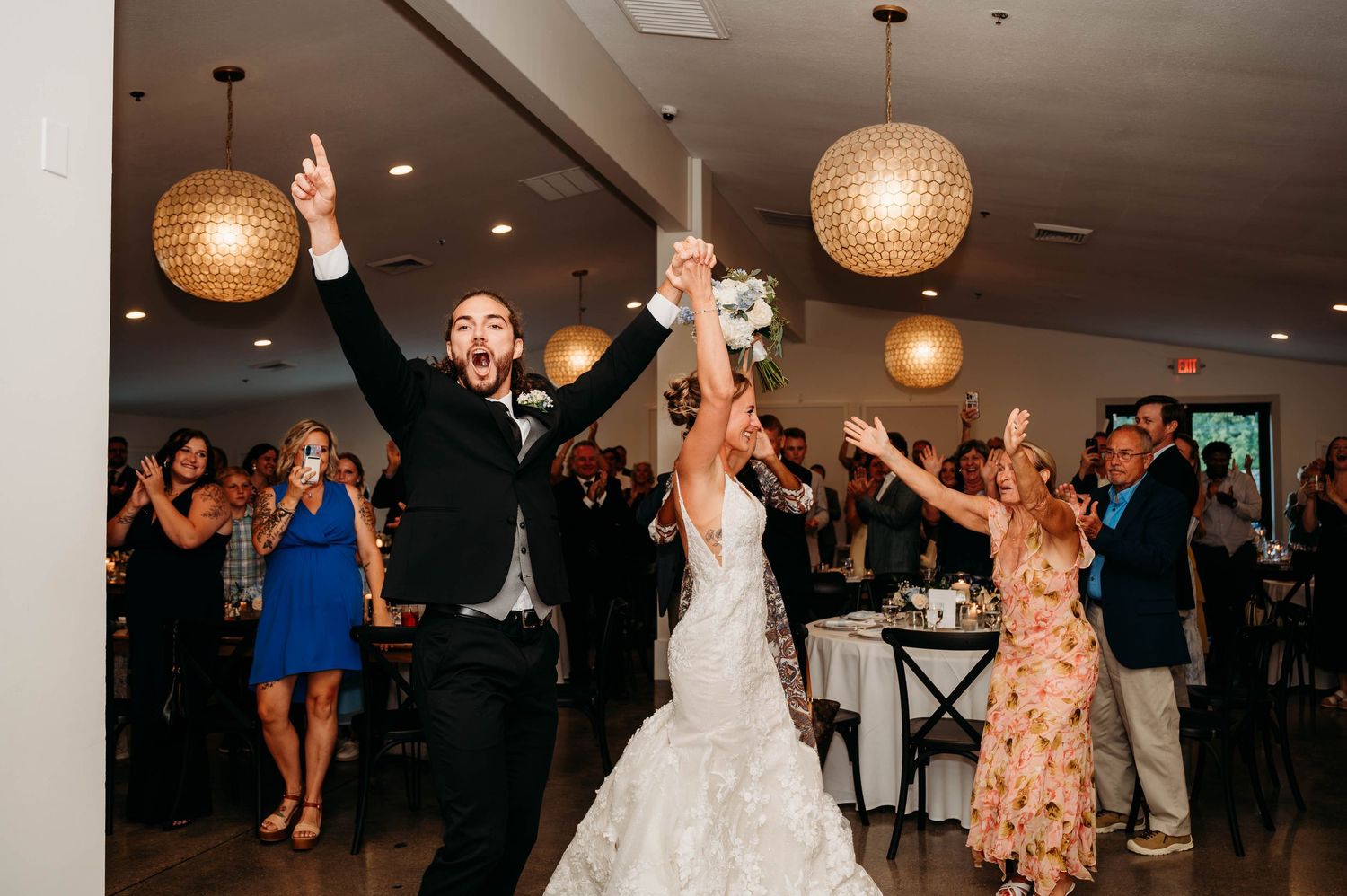 Joyful newlyweds raise their arms in celebration as they enter their elegant wedding reception hall with guests cheering.