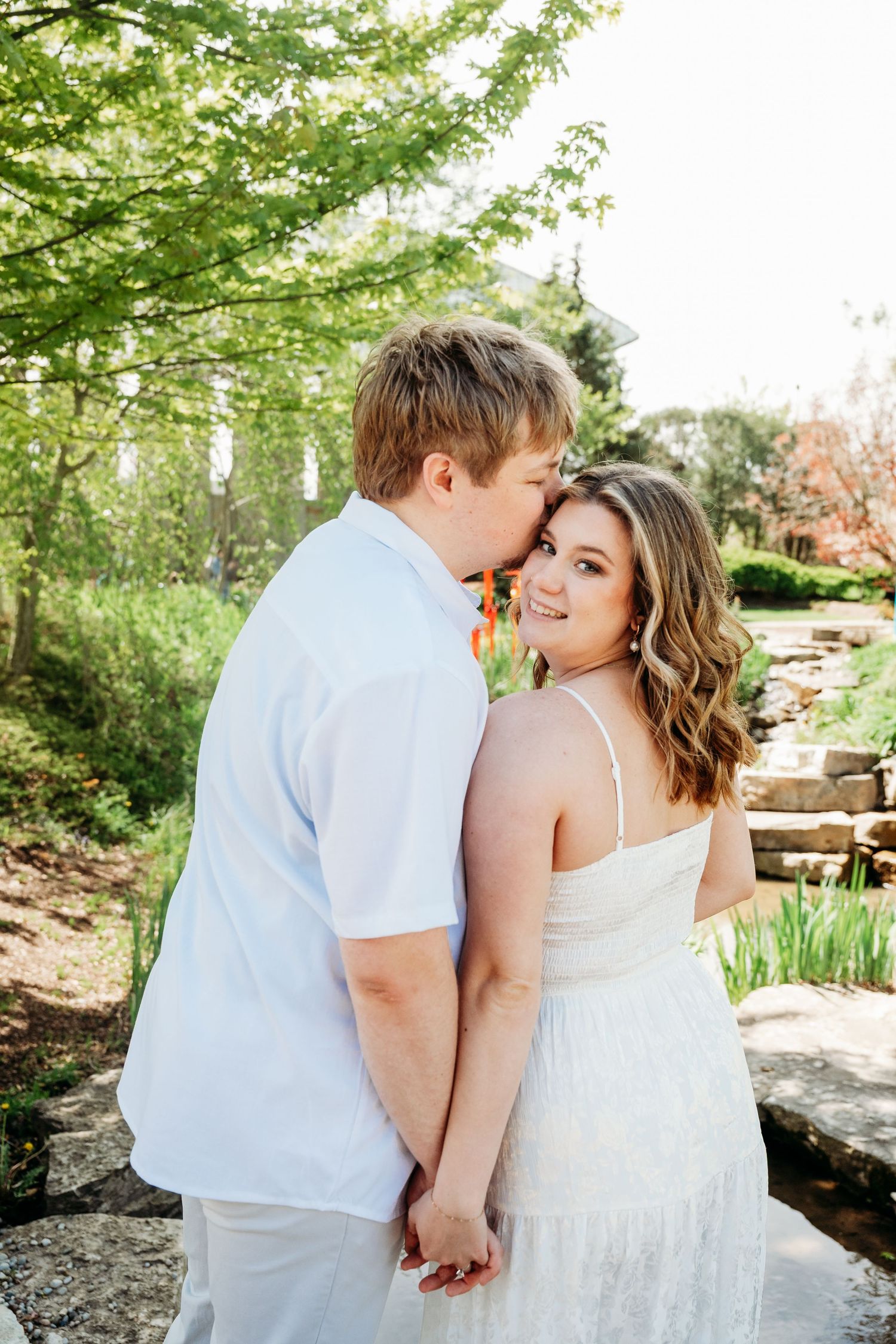 A couple in white outfits holds hands outdoors, the man kissing the woman's cheek as she smiles at the camera.