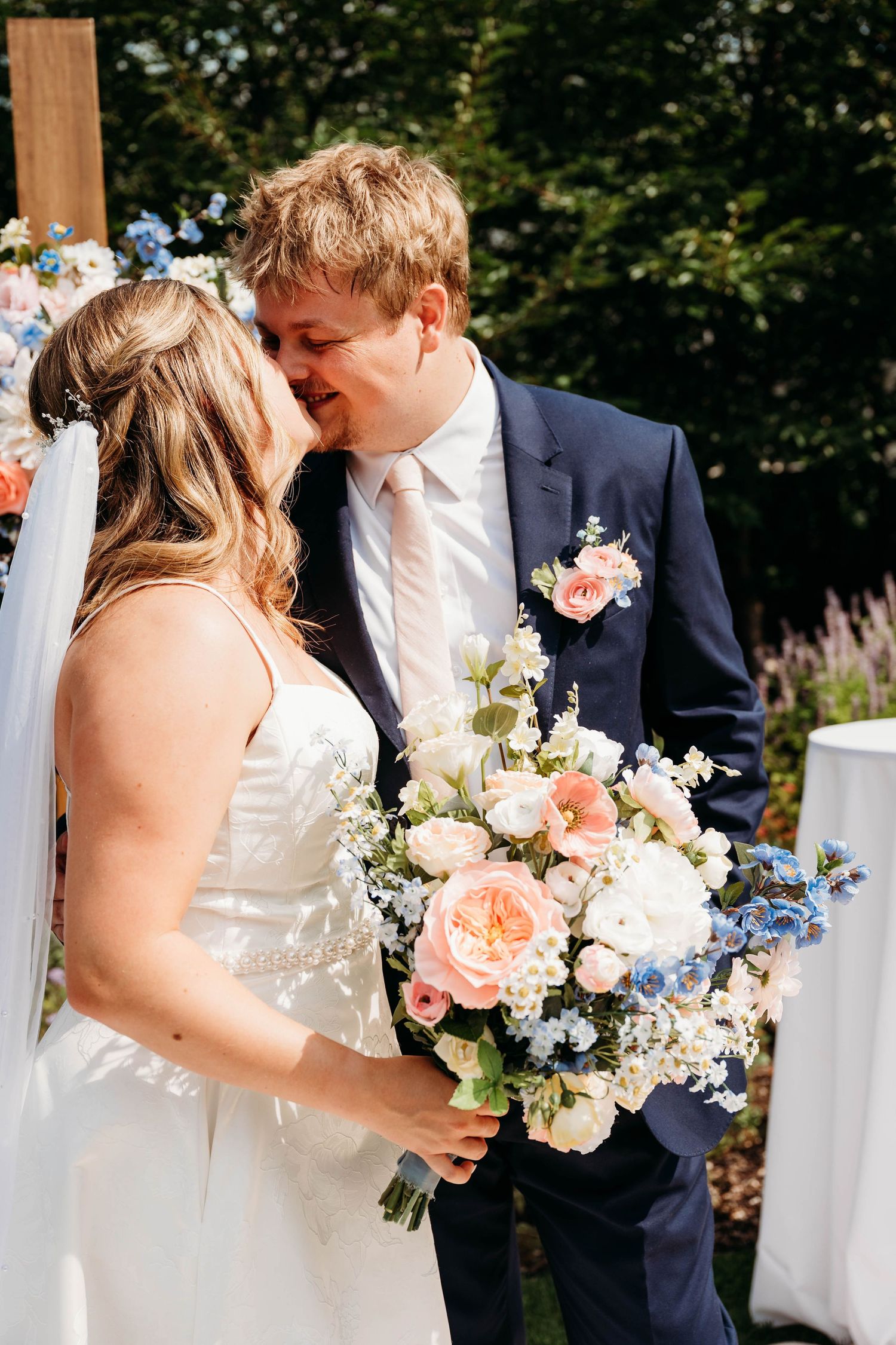Bride and groom share first kiss at outdoor wedding ceremony, bride holding pink and blue floral bouquet.