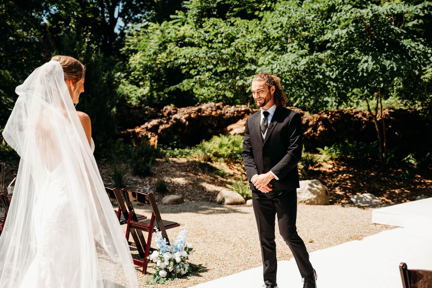 Groom smiles seeing bride in white gown and veil during outdoor wedding first look ceremony.