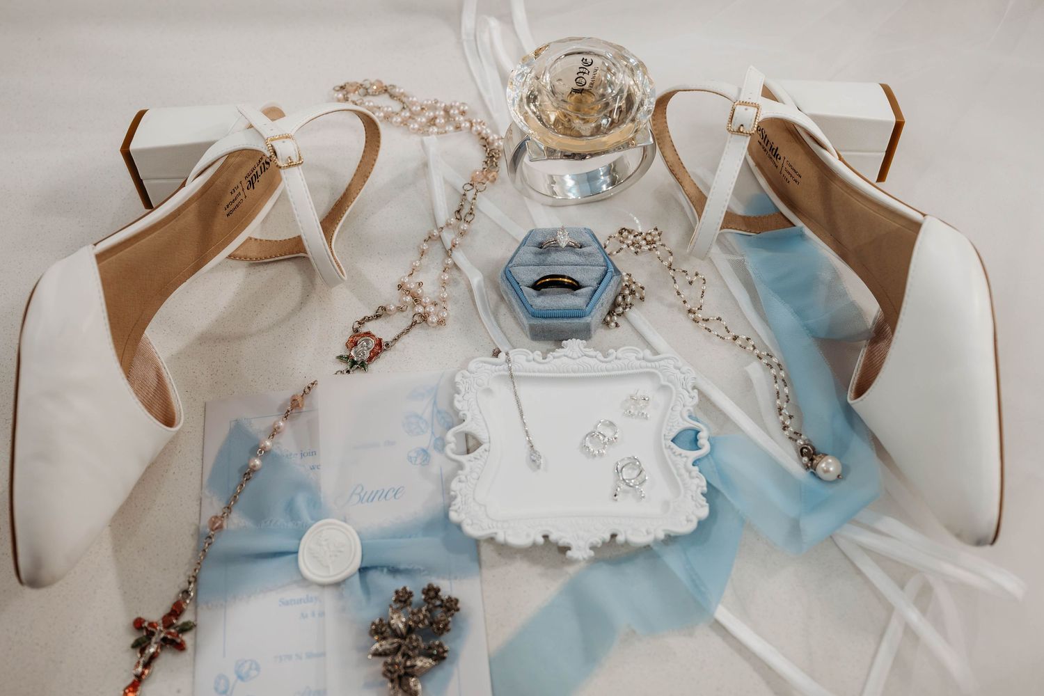 Elegant bridal flat lay with white heels, blue ribbon, rosary, ring box, and delicate jewelry on soft fabric.