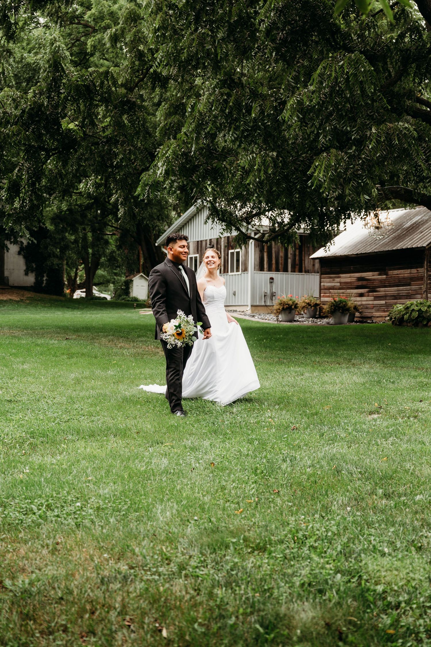 Bride and groom walking hand in hand on green lawn with bouquet, trees and rustic barn in background.