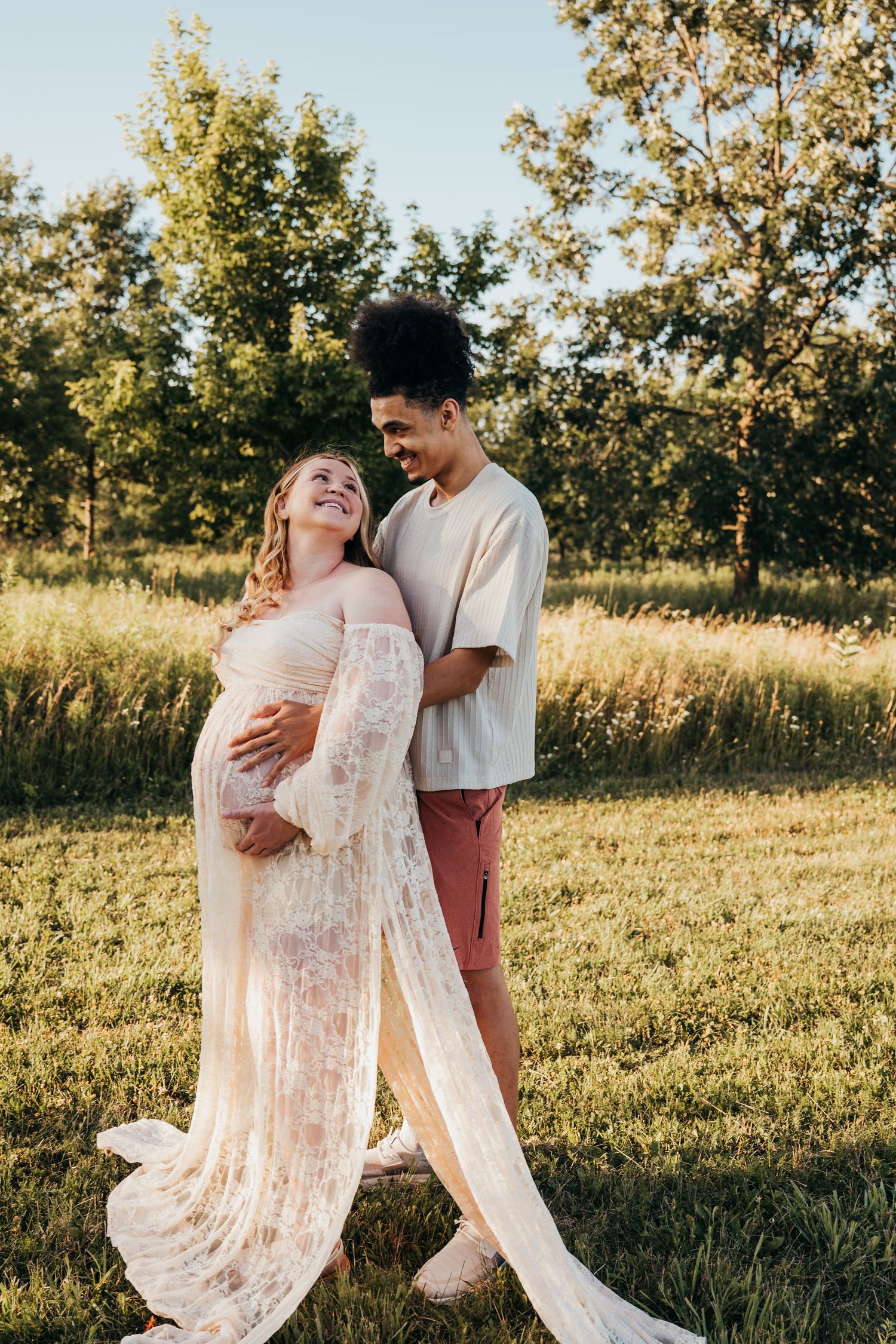 Expecting couple embraces in a sunset field, with the pregnant partner wearing a flowing lace dress.