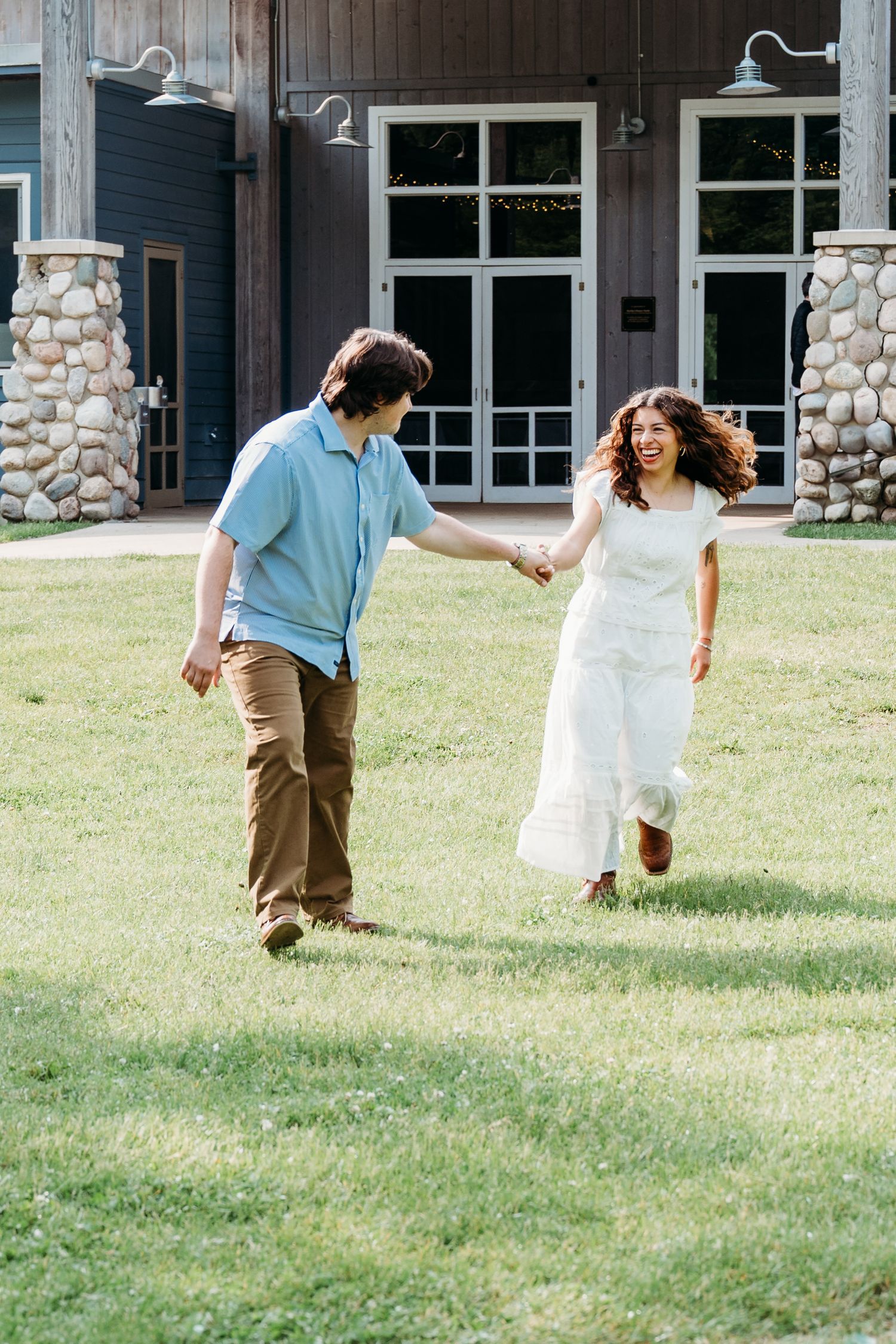 Couple running and holding hands on a grassy lawn in front of a building with stone pillars and large windows.