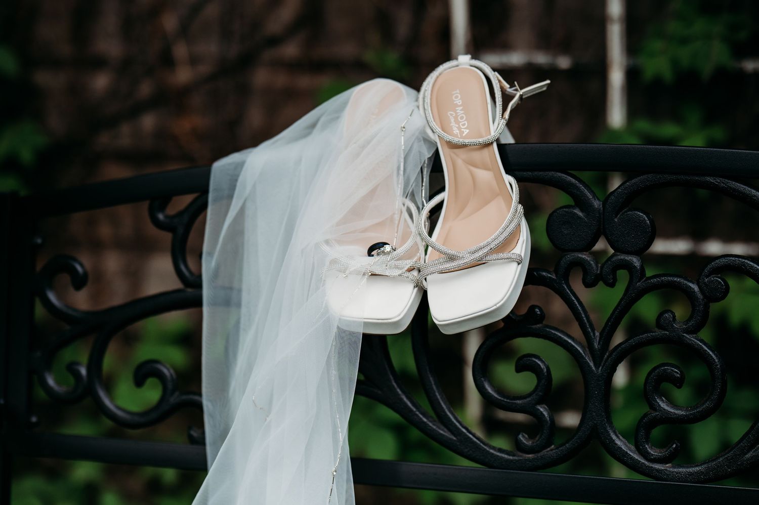 White wedding shoes displayed on decorative iron railing with lace veil draped nearby at outdoor venue.