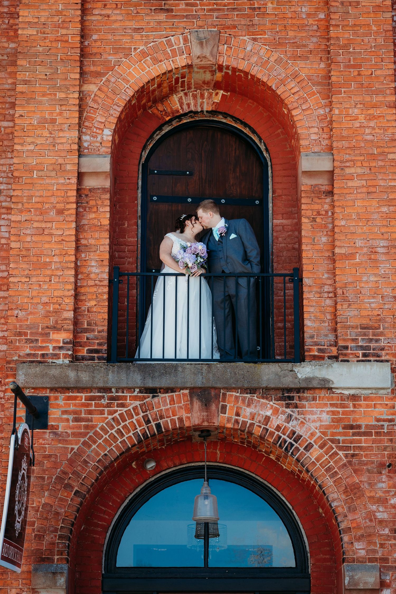 A romantic wedding couple embraces in an arched brick doorway of a historic building.