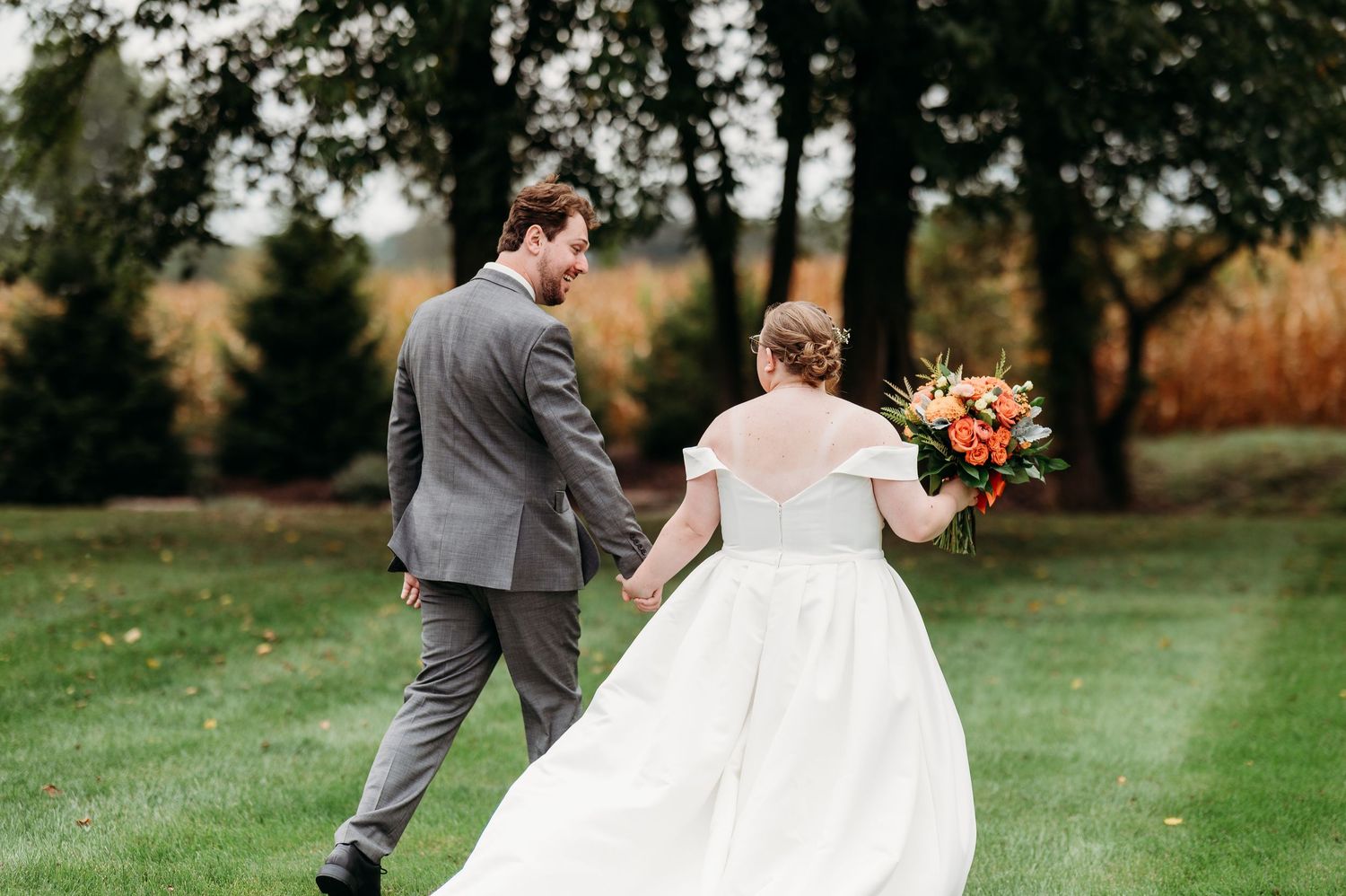 A couple in wedding attire holds hands while walking through a lush green field during an autumn day.