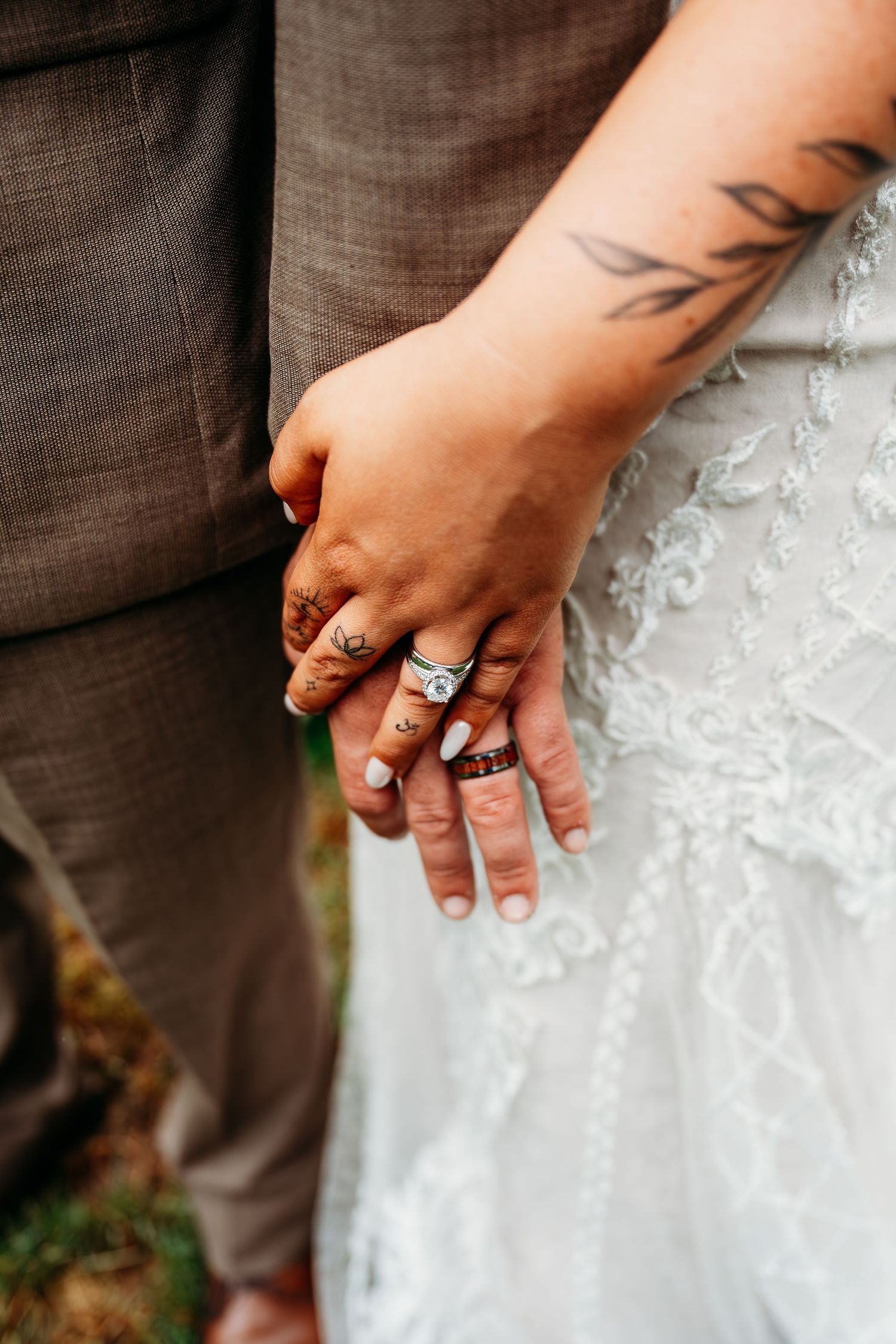 Close-up of bride and groom holding hands showing wedding rings and tattoos against a lace wedding gown.