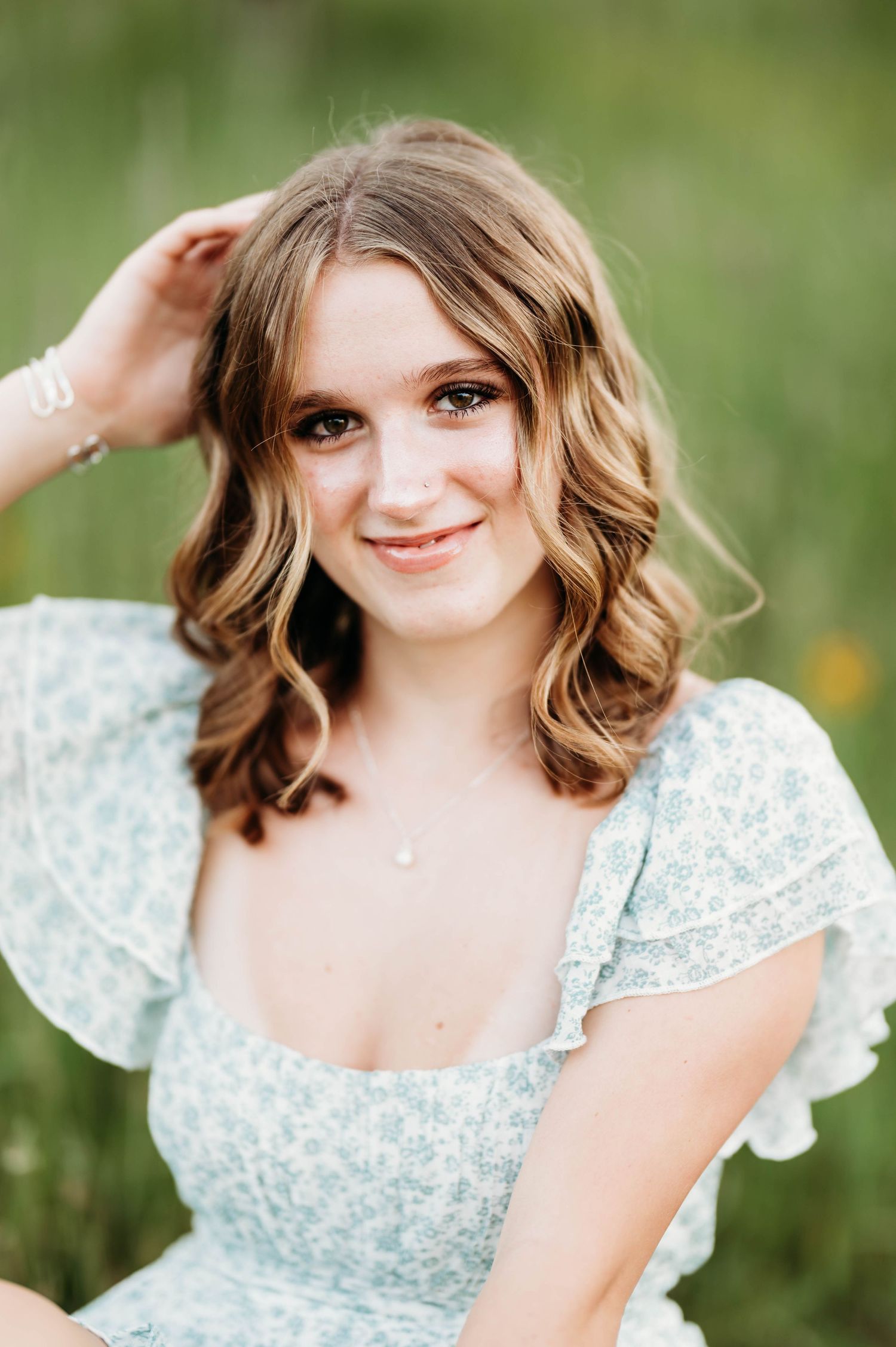 Young woman in light blue floral dress smiling outdoors in green field during senior portrait session.