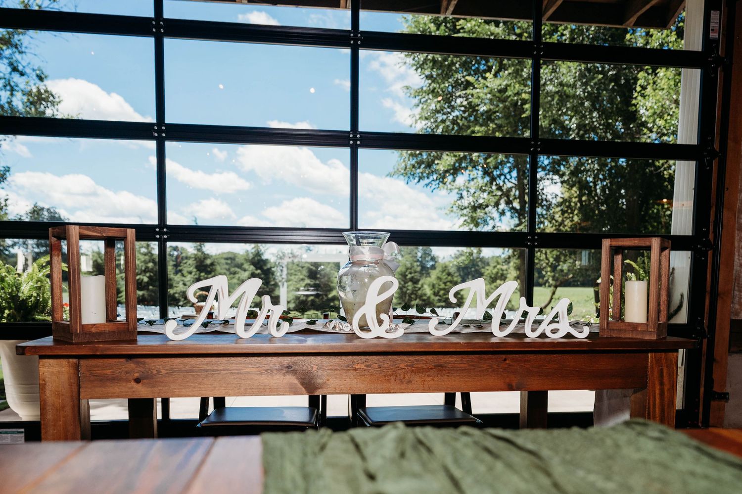 Rustic wooden sweetheart table with Mr. & Mrs. sign, framed photos, and mason jars in front of large windows.