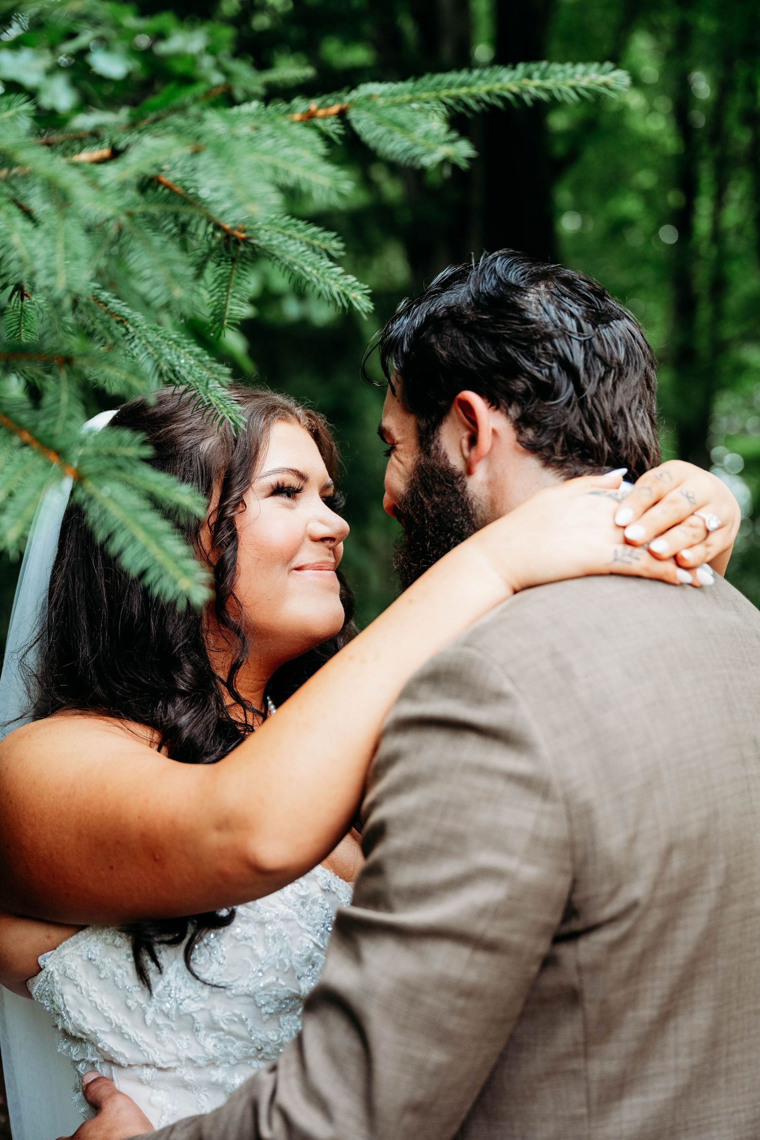 Bride smiles at bearded groom while embracing outdoors under lush green pine tree branches.